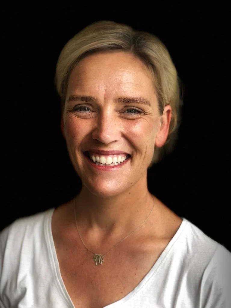 Smiling woman with short blonde hair wearing a white shirt and a necklace, against a dark background.