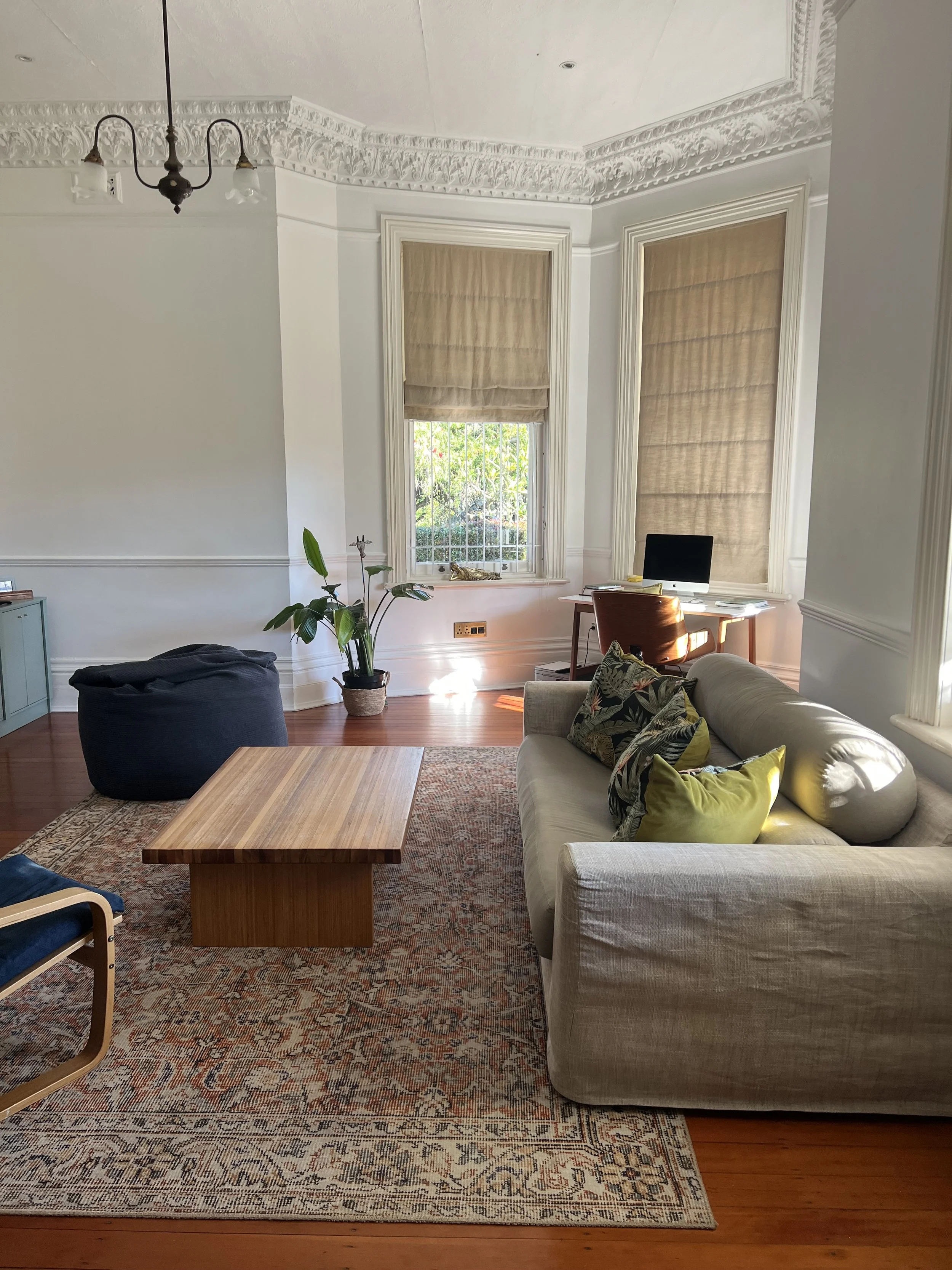 Living room with a beige sofa, pillows, a wooden coffee table, a patterned area rug, a potted plant, a window with blinds, a desk with a computer, and a chandelier with two lights.