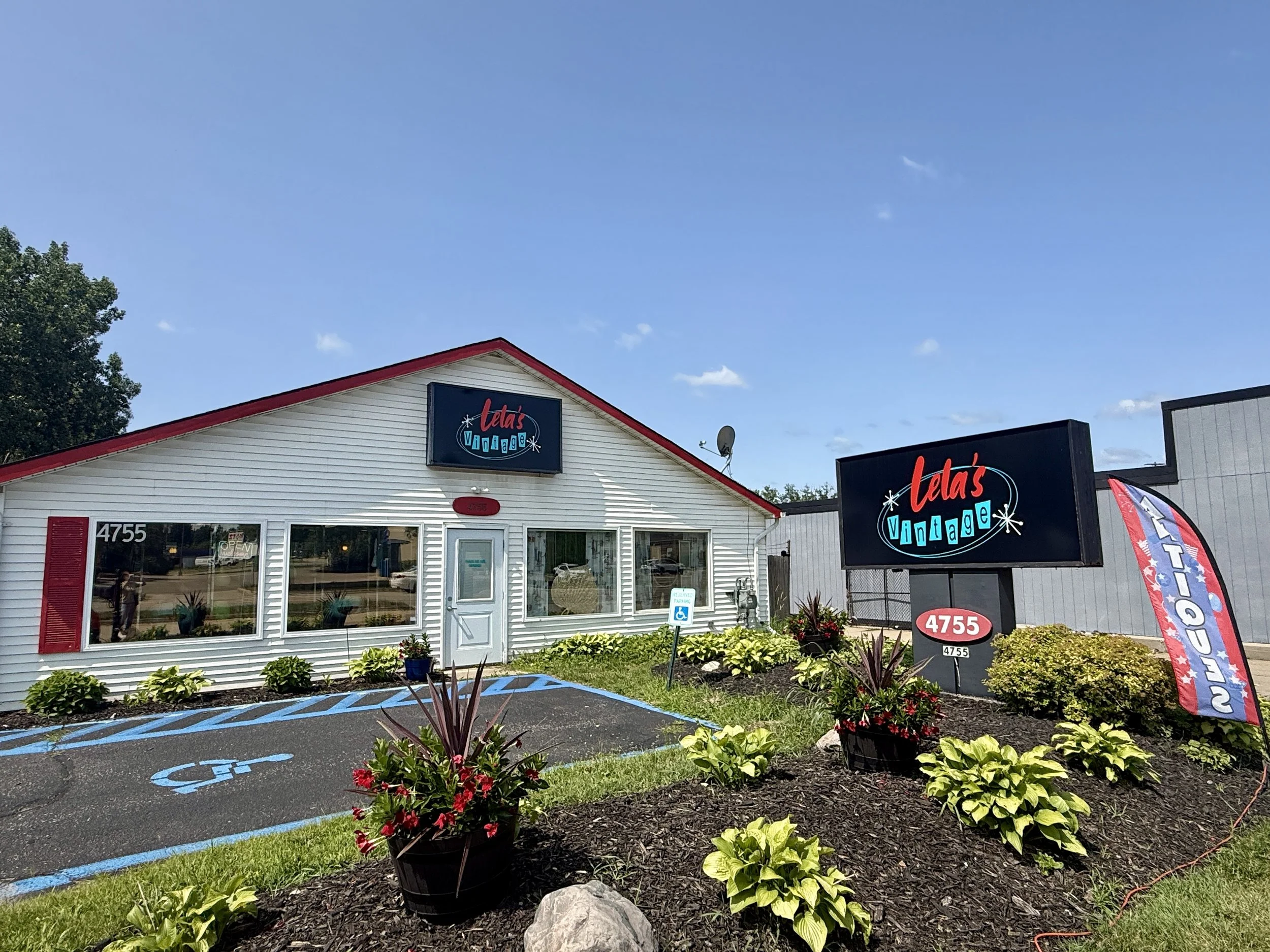 Front view of Lela's Vintage store with signage, parking spaces including accessible spot, and landscaping with flowers and plants under a blue sky.