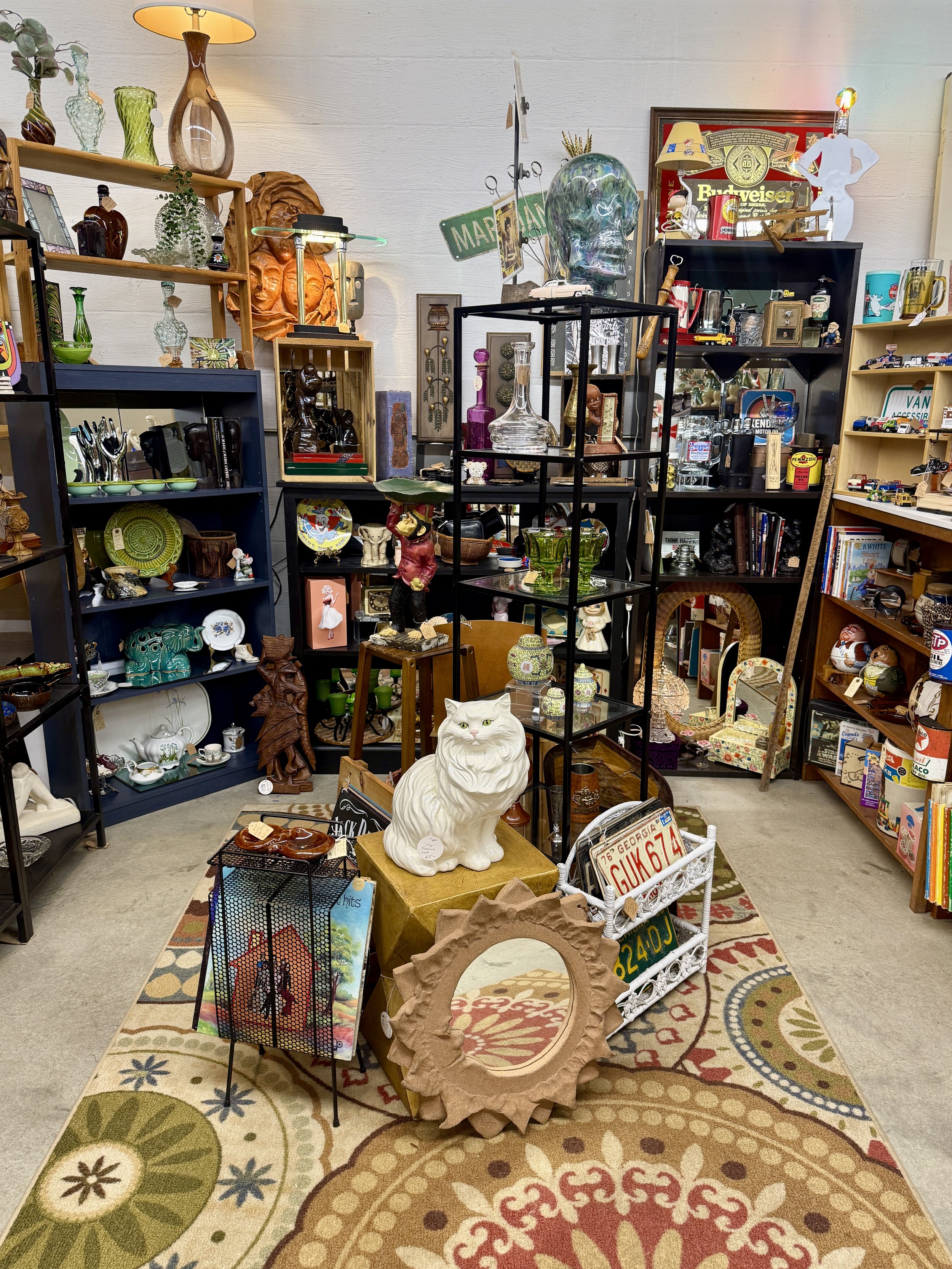 A thrift store display with various decorative items including vases, lamps, figurines, glassware, and collectibles arranged on shelves and tables. The floor has a patterned rug and miscellaneous objects are scattered throughout the scene.