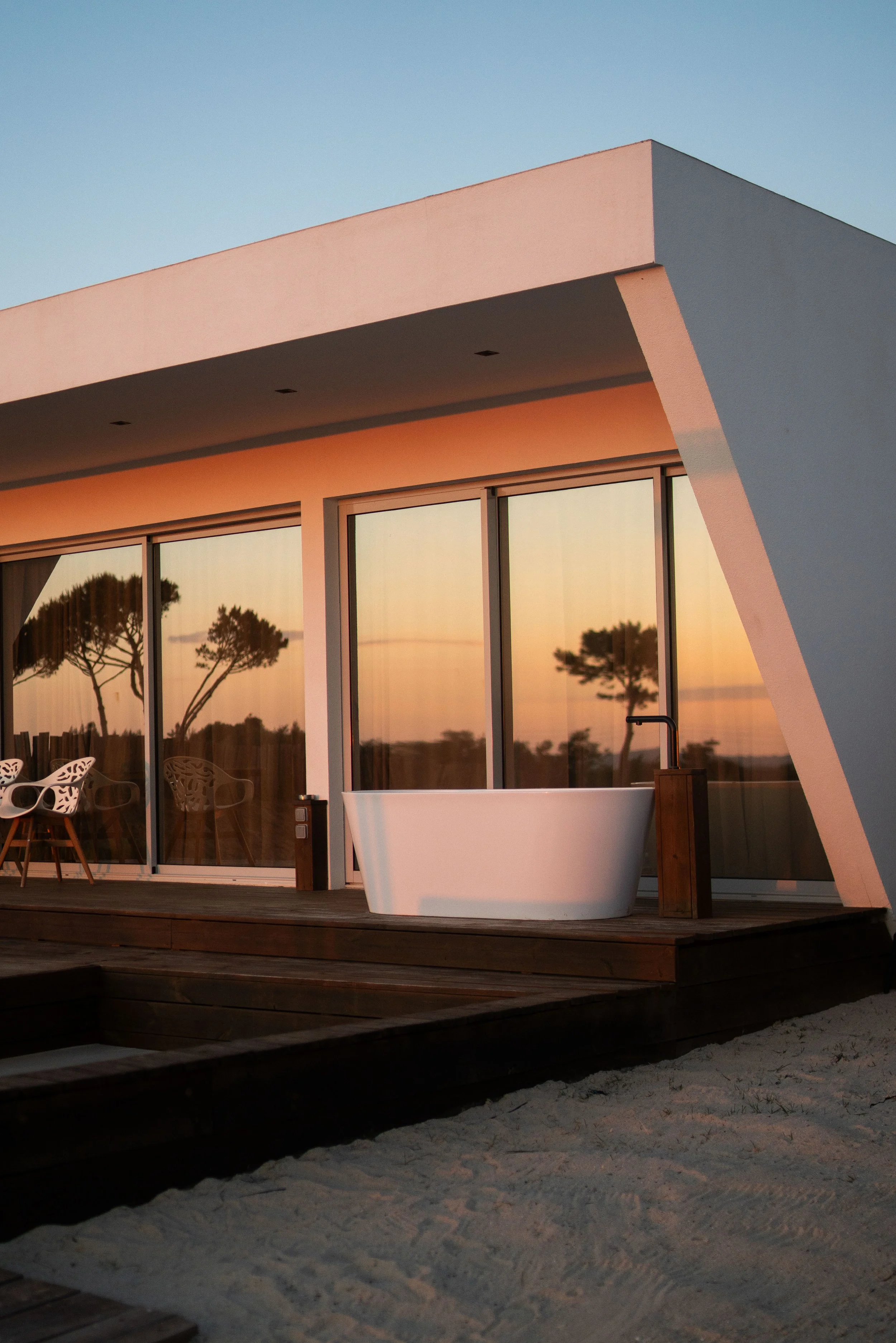 Modern house with large glass windows reflecting a sunset sky and trees, featuring a wooden deck with outdoor seating and a freestanding white bathtub on a sandy ground.