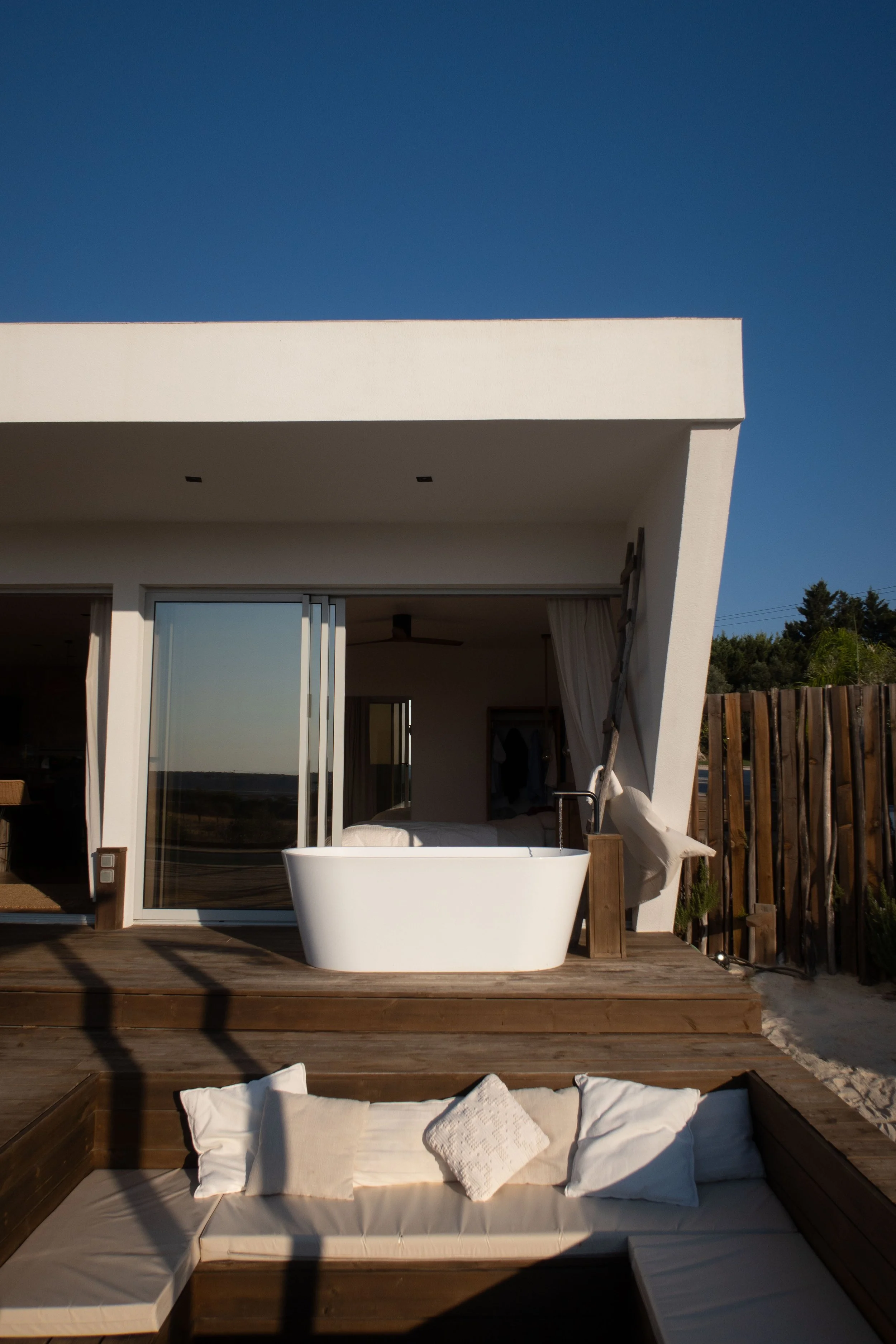 Outdoor patio with a white modern building, sliding glass door, bathtub, and cushioned seating area with pillows.