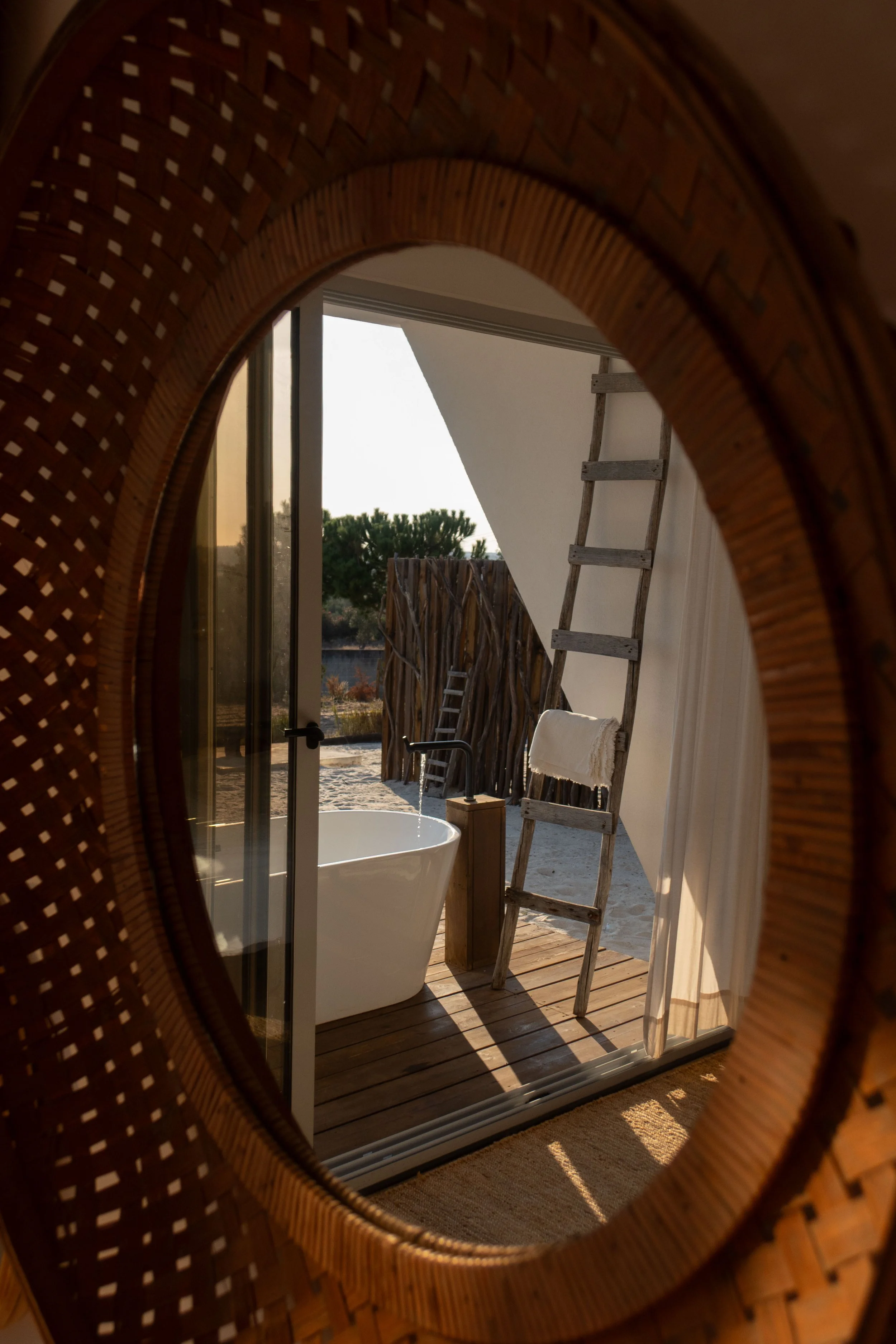 Looking through a mirror at a balcony with a freestanding bathtub, ladder, and outdoor scenery