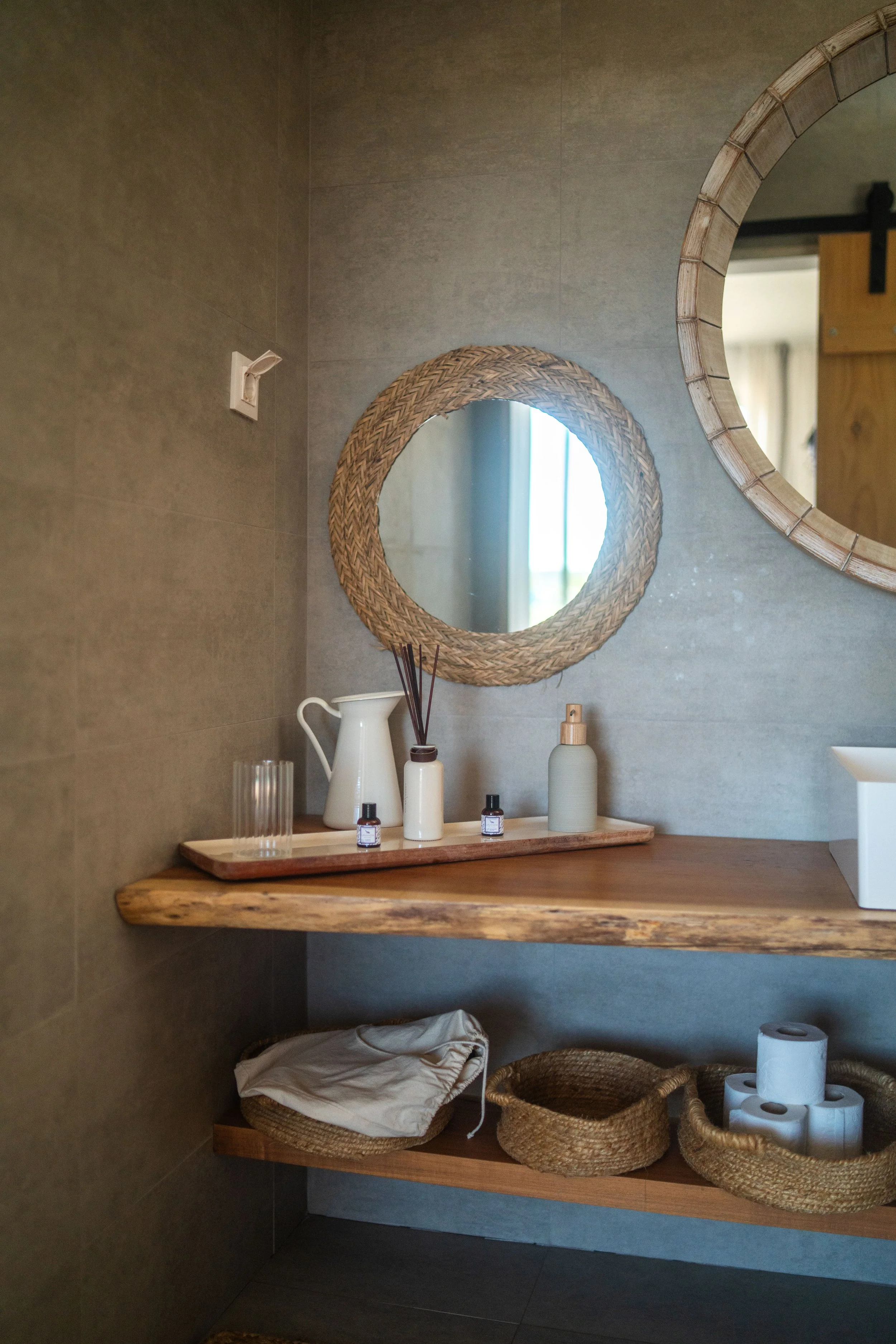 Decorative bathroom corner with two round mirrors, wooden shelves holding bottles, a white pitcher, glass, crocheted baskets, toilet paper rolls, and a wicker basket on the bottom shelf.
