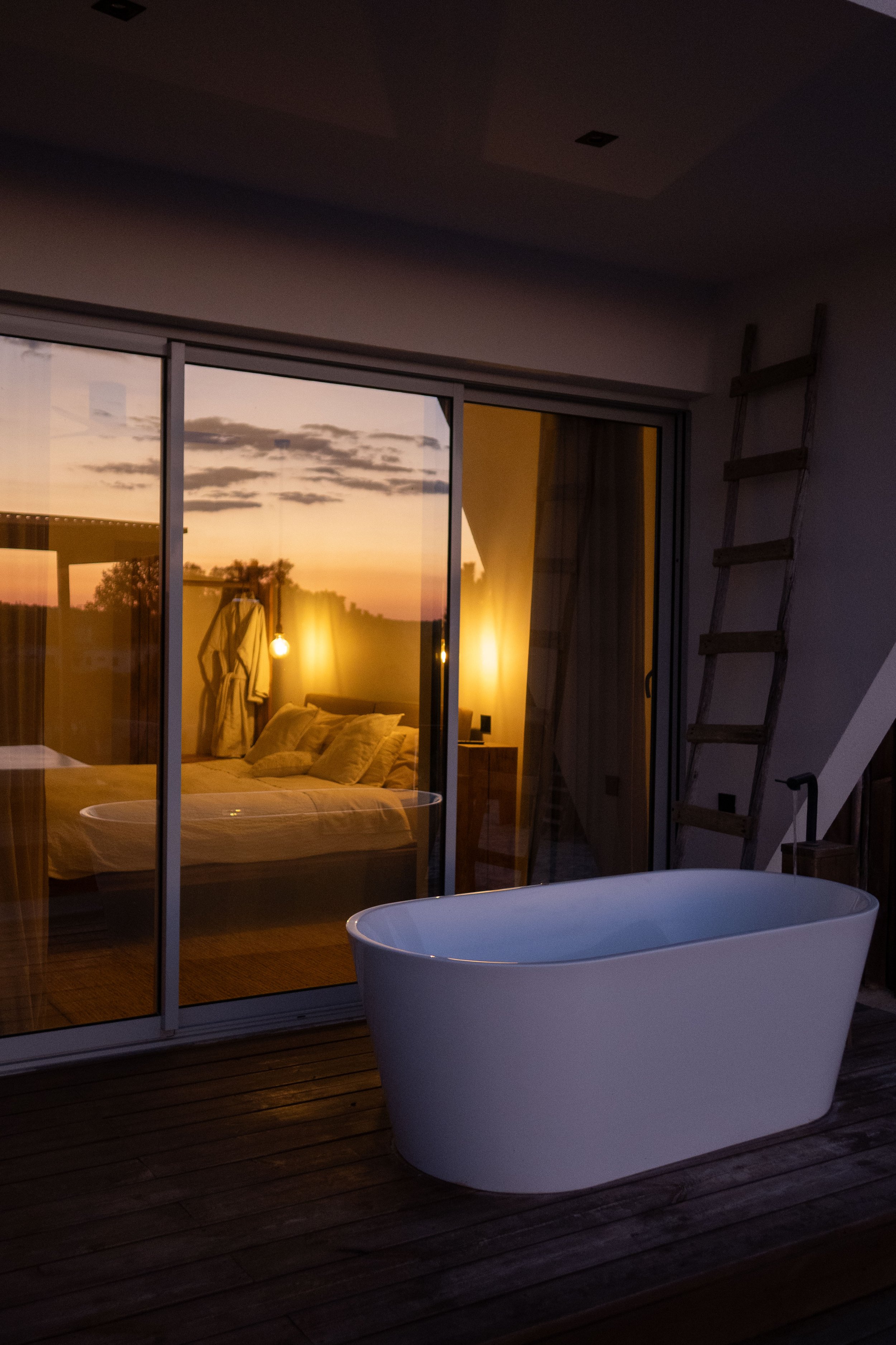 A bedroom seen through a glass sliding door with a sunset sky outside, a bed with pillows and a robe hanging on the back of a chair inside, illuminated by warm yellow lights, with a bathtub on a wooden deck outside.