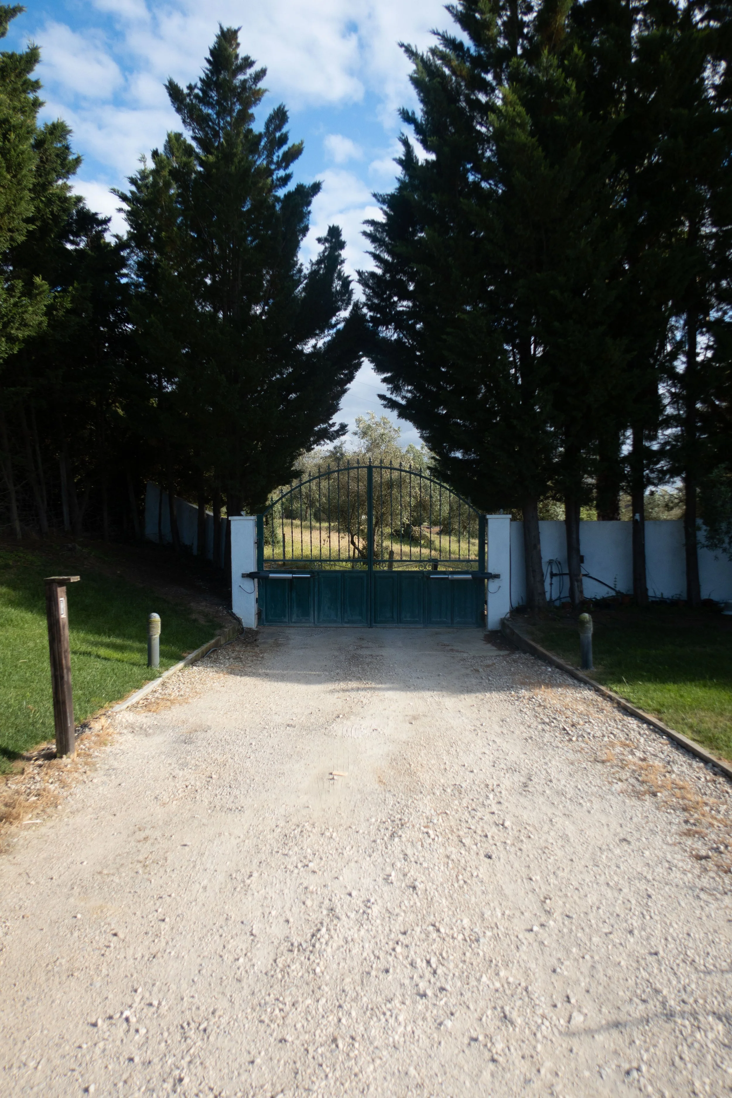 A gravel driveway leading to a black metal gate, flanked by white pillars, surrounded by tall green trees on both sides, with a partly cloudy sky overhead.