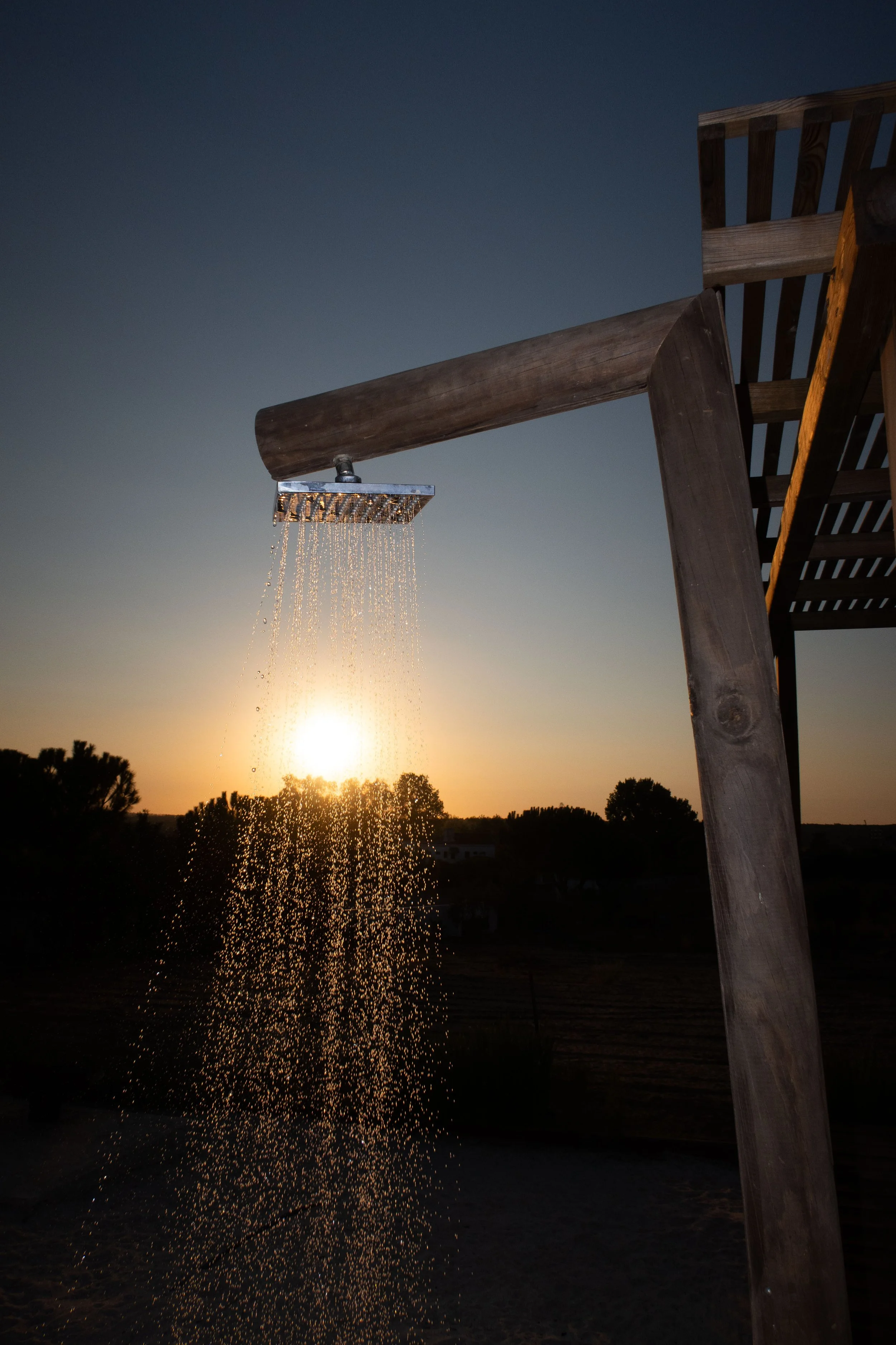 Outside shower with water flowing from a large wooden showerhead at sunset, trees and a distant horizon visible in the background.