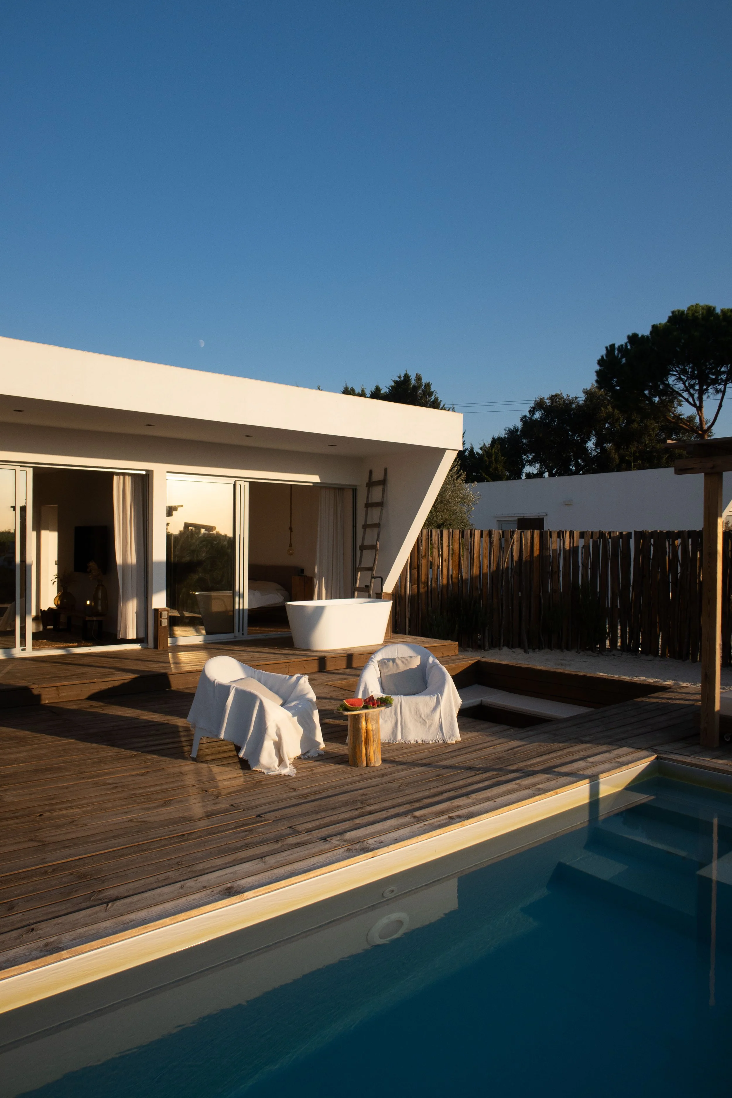 Modern house with large glass sliding doors, outdoor wooden deck, outdoor seating with white chairs and small table, pool with steps, private backyard with wooden fence, and clear blue sky with visible moon.