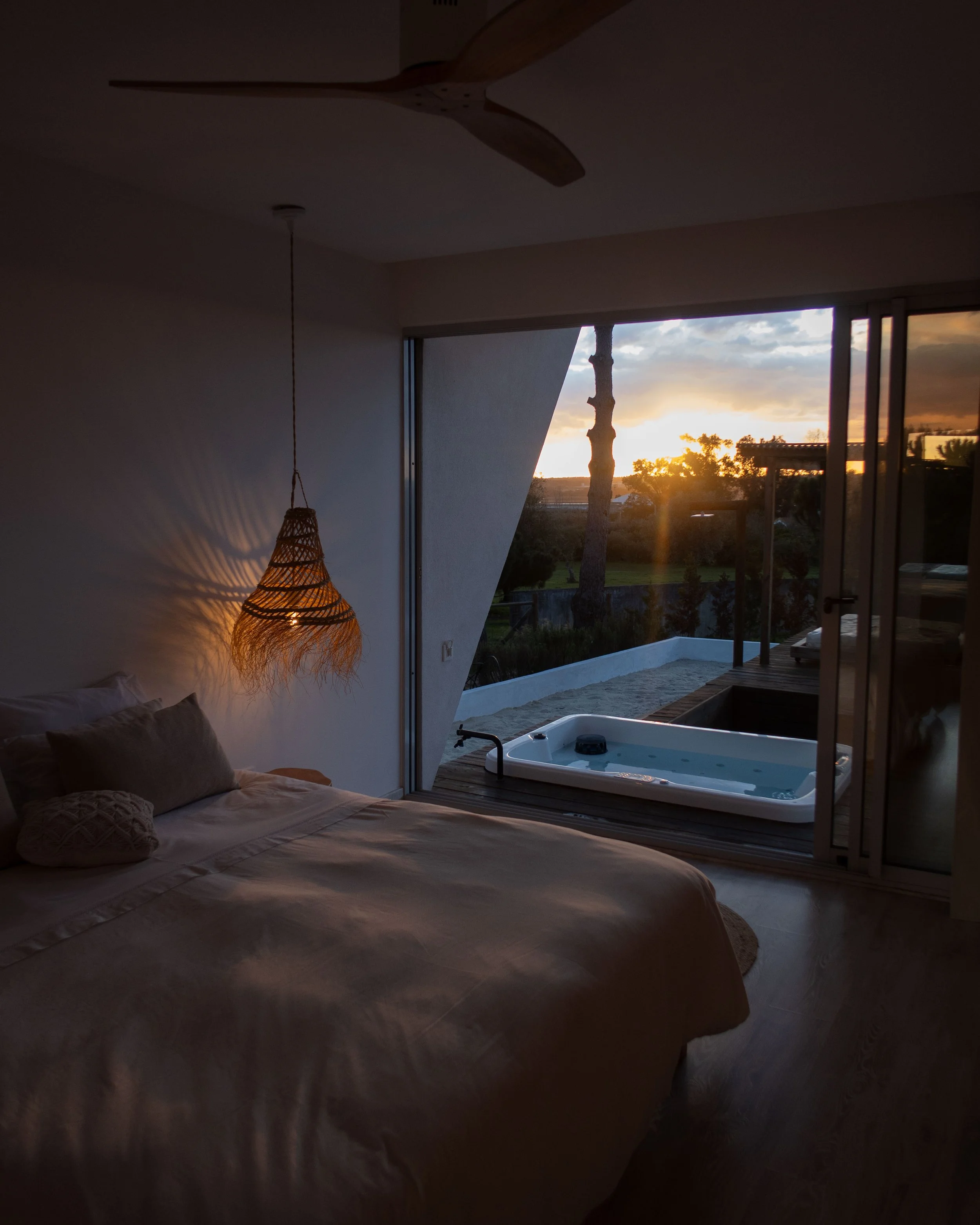 Bedroom with large windows showing sunset and outdoor scenery, bed with pillows, hanging woven lamp, jacuzzi on the balcony.