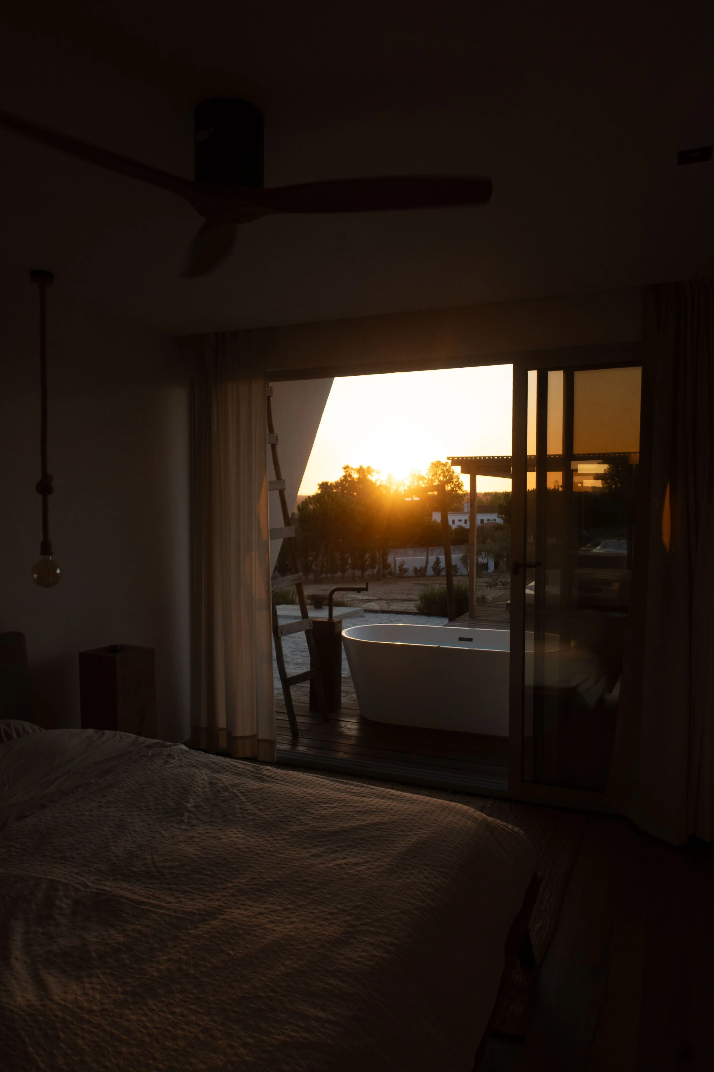A bedroom with a large bed, a sliding glass door, and an outdoor hot tub, with the sun setting in the background.