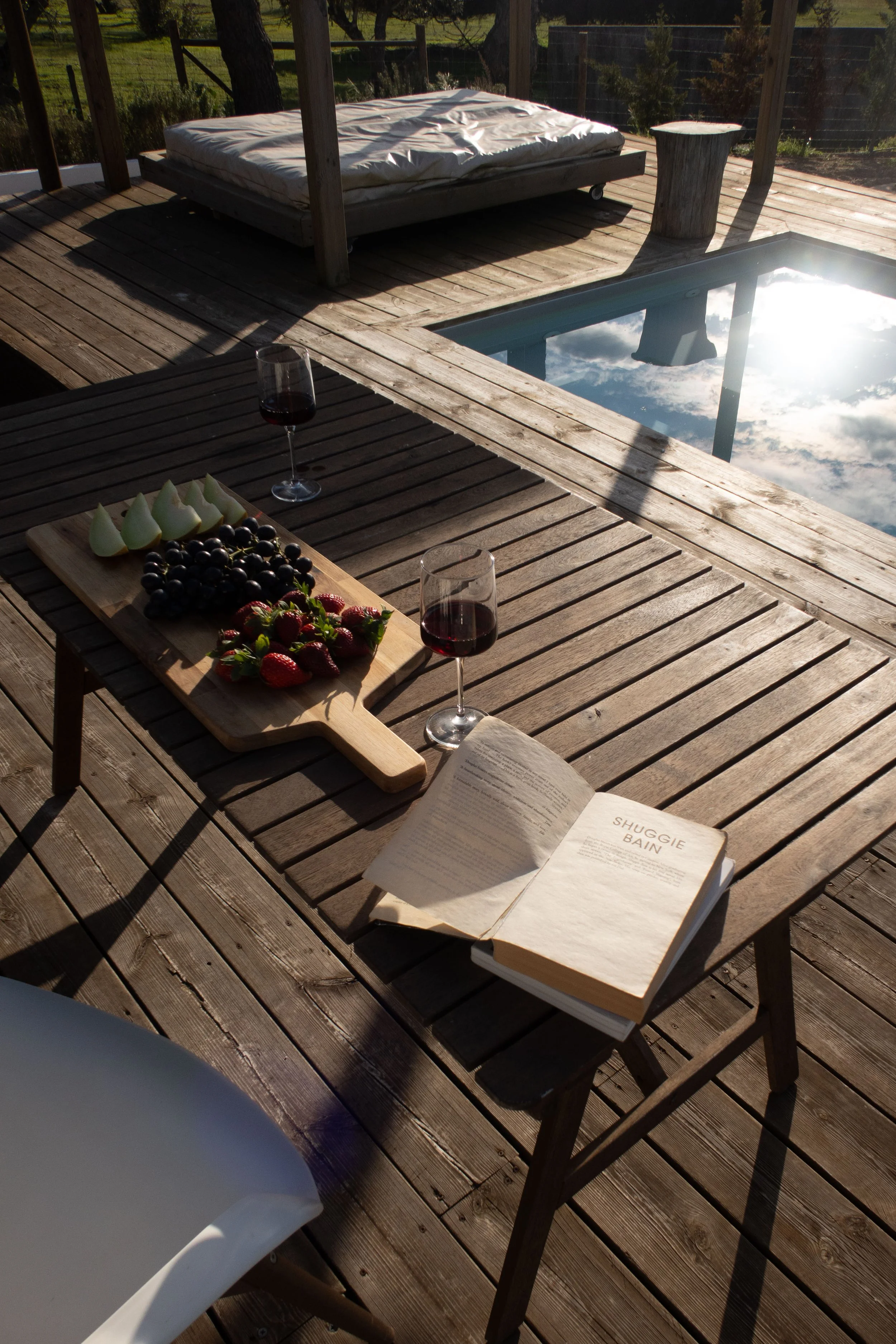 An outdoor wooden deck with a hot tub reflecting the sky, a mattress on a wooden platform, a tree stump side table, a wooden table with a glass of red wine, a cheese and fruit platter, and an open book titled 'Shuggie Bain'.