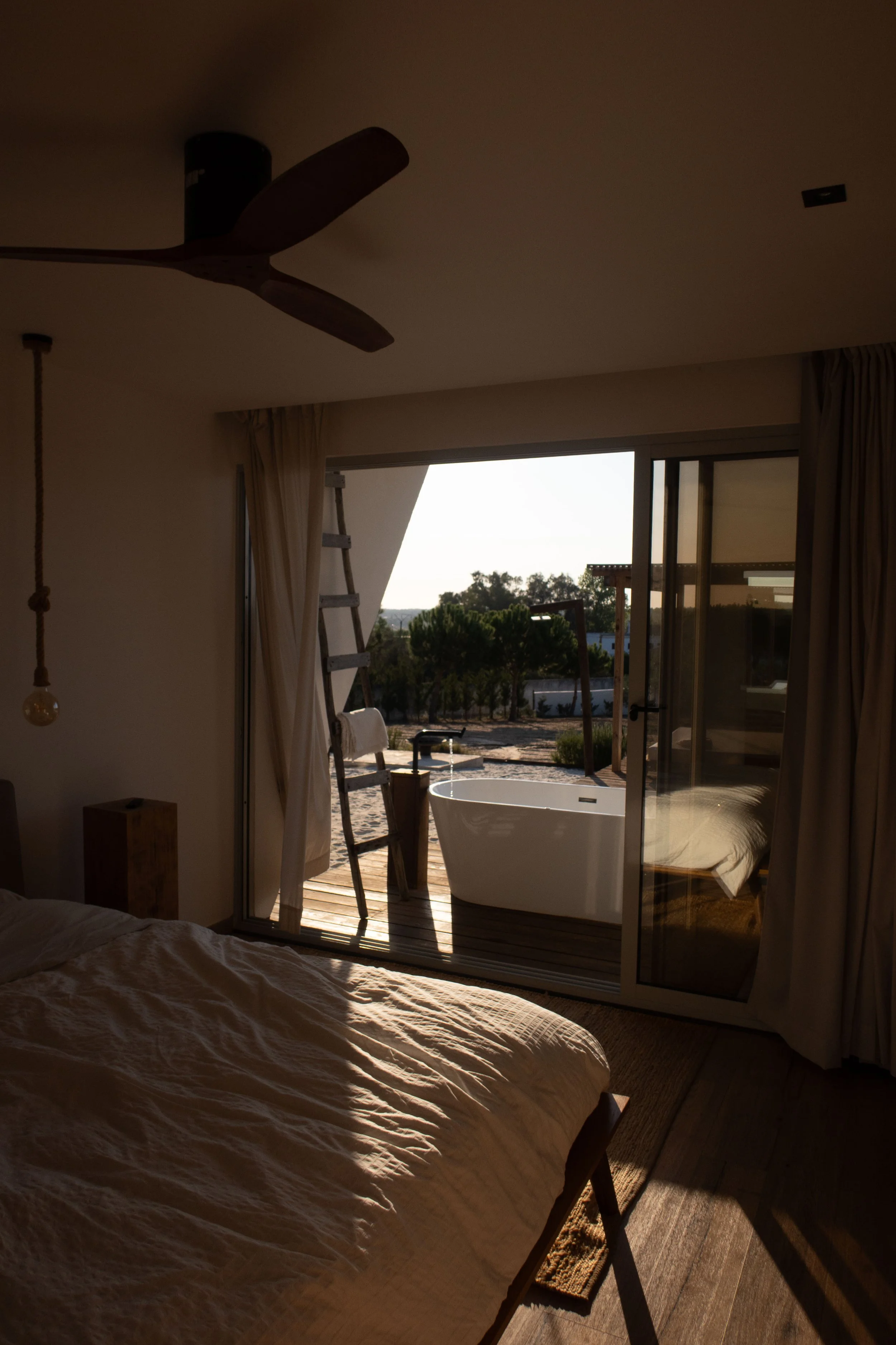 Bedroom with bed, curtains, ladder, outdoor bathtub on balcony, and sliding glass door leading outside, with trees and sky visible.