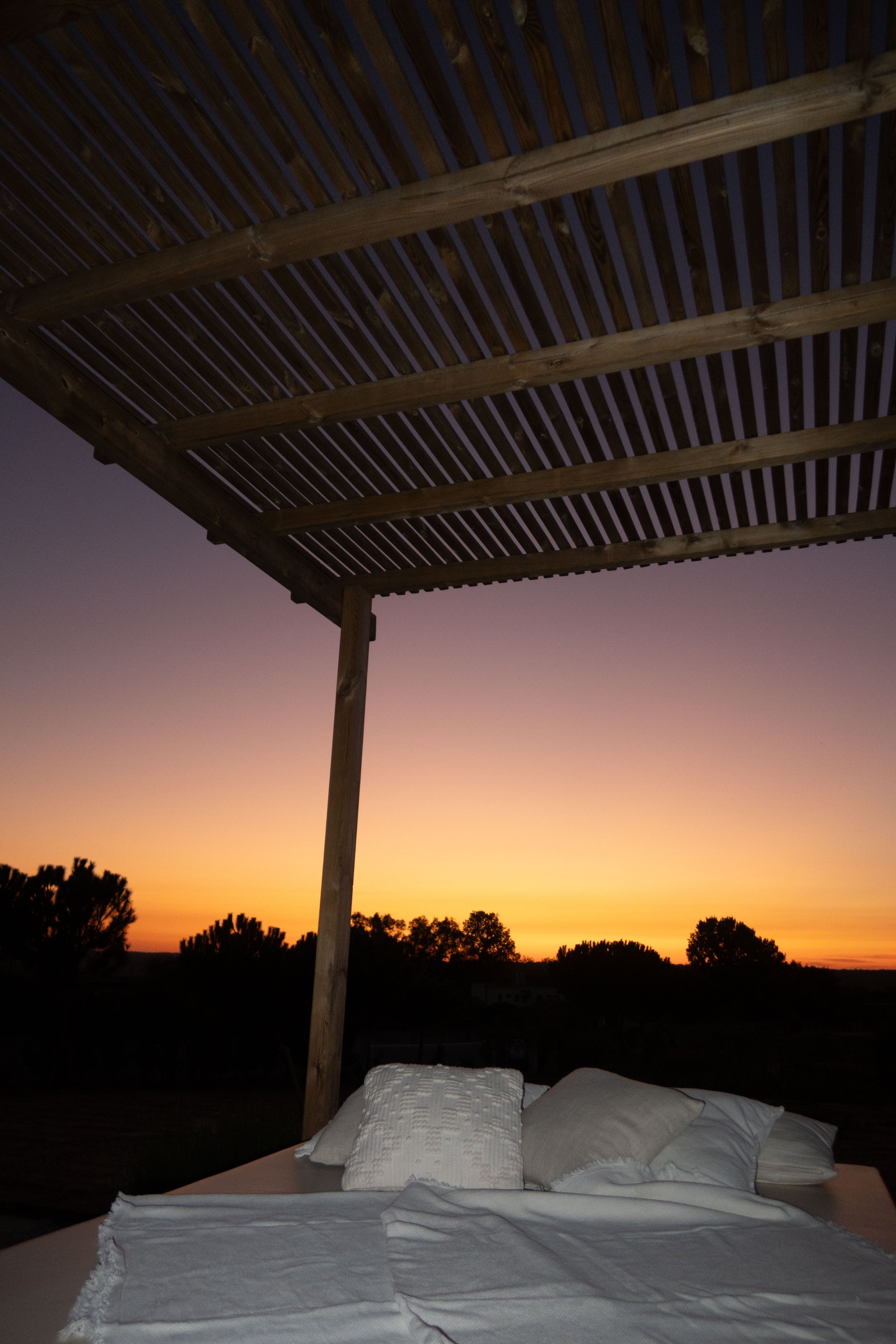 A bed with white pillows and sheets under a wooden canopy structure at sunset, with trees silhouetted in the distance.