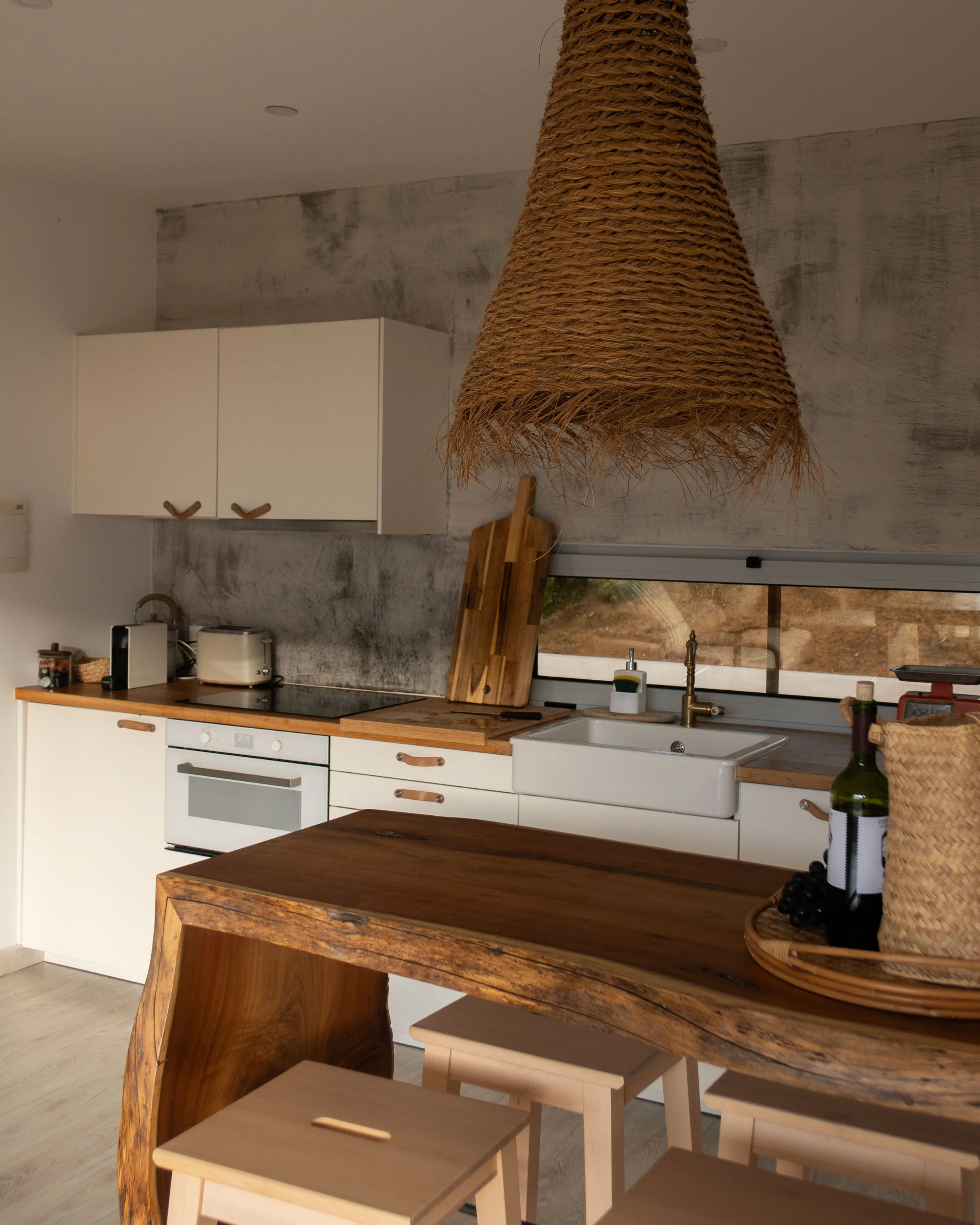 A modern kitchen with white cabinets, wooden countertops, and a large wooden dining table. The kitchen has a white sink with a gold faucet, bottles, and a wine bottle on the table. There is a wicker pendant light hanging from the ceiling and a window
