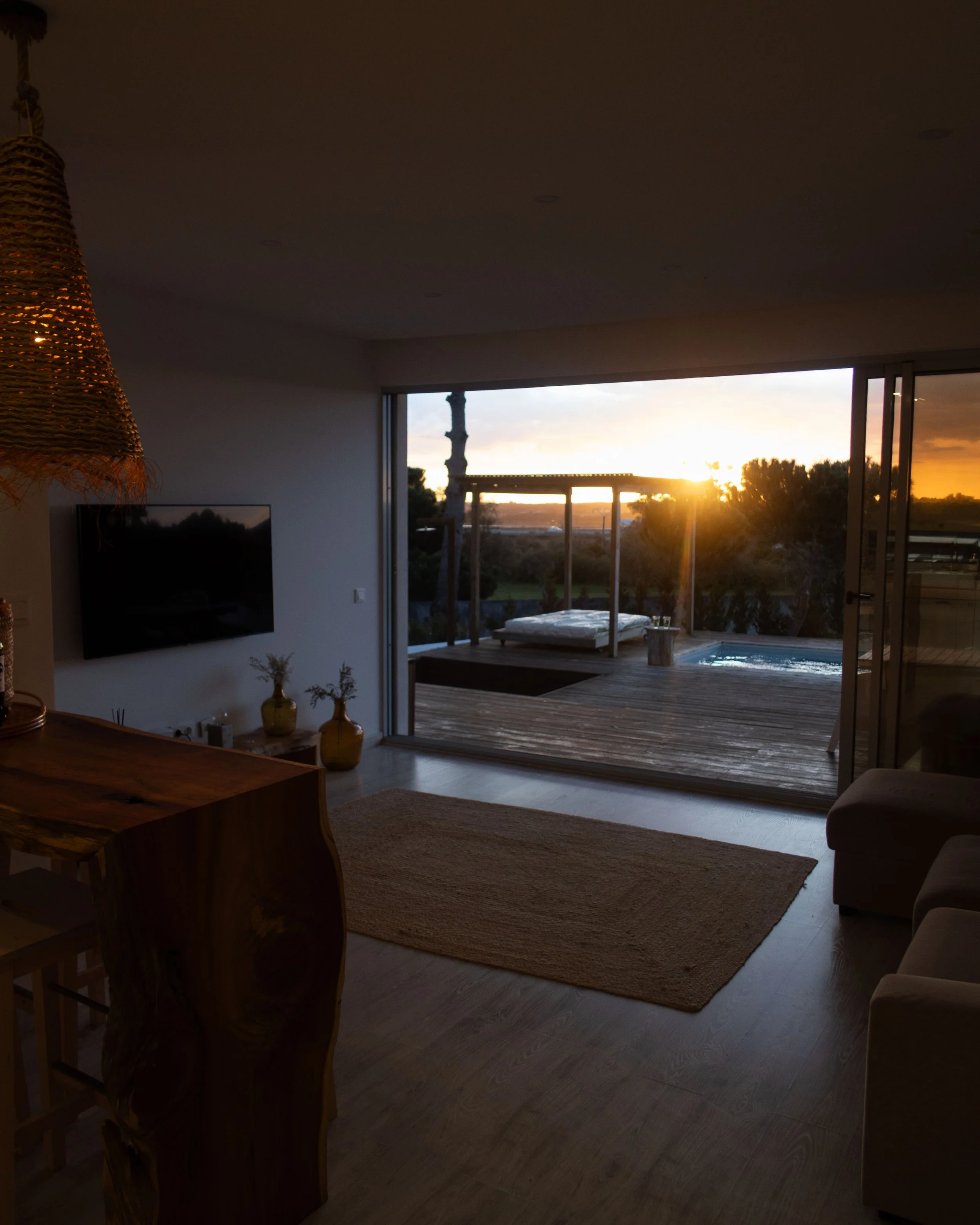 Interior of a modern living room with large sliding glass doors opening to an outdoor patio with a hot tub, wooden deck, and sunset view outside.