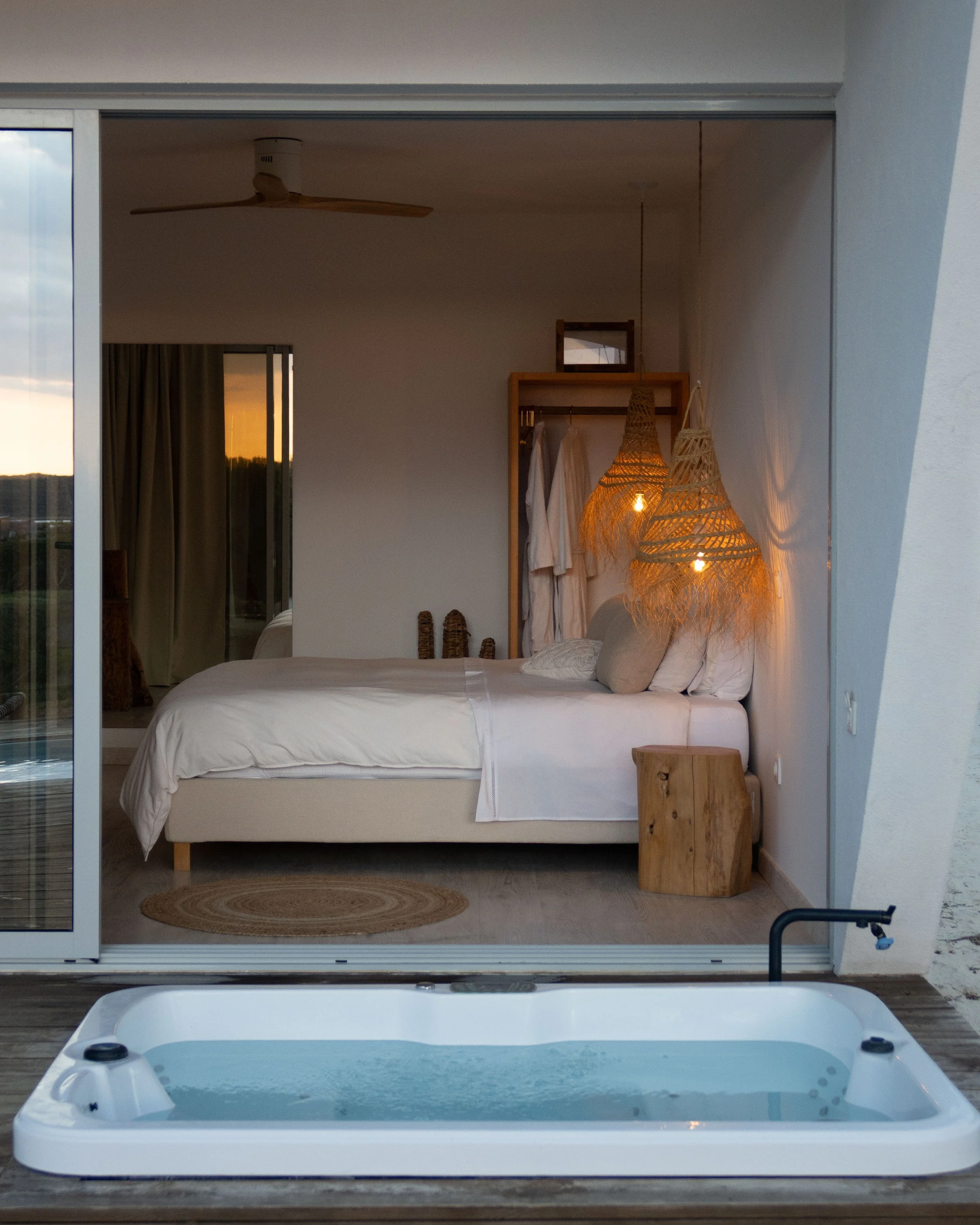 A view from outside a building showing a hot tub on a deck, with a bedroom visible through open sliding glass doors. The room has a bed with white bedding, a hanging wicker lamp, a wooden stool, and a clothing rack with white robes. A ceiling fan is 