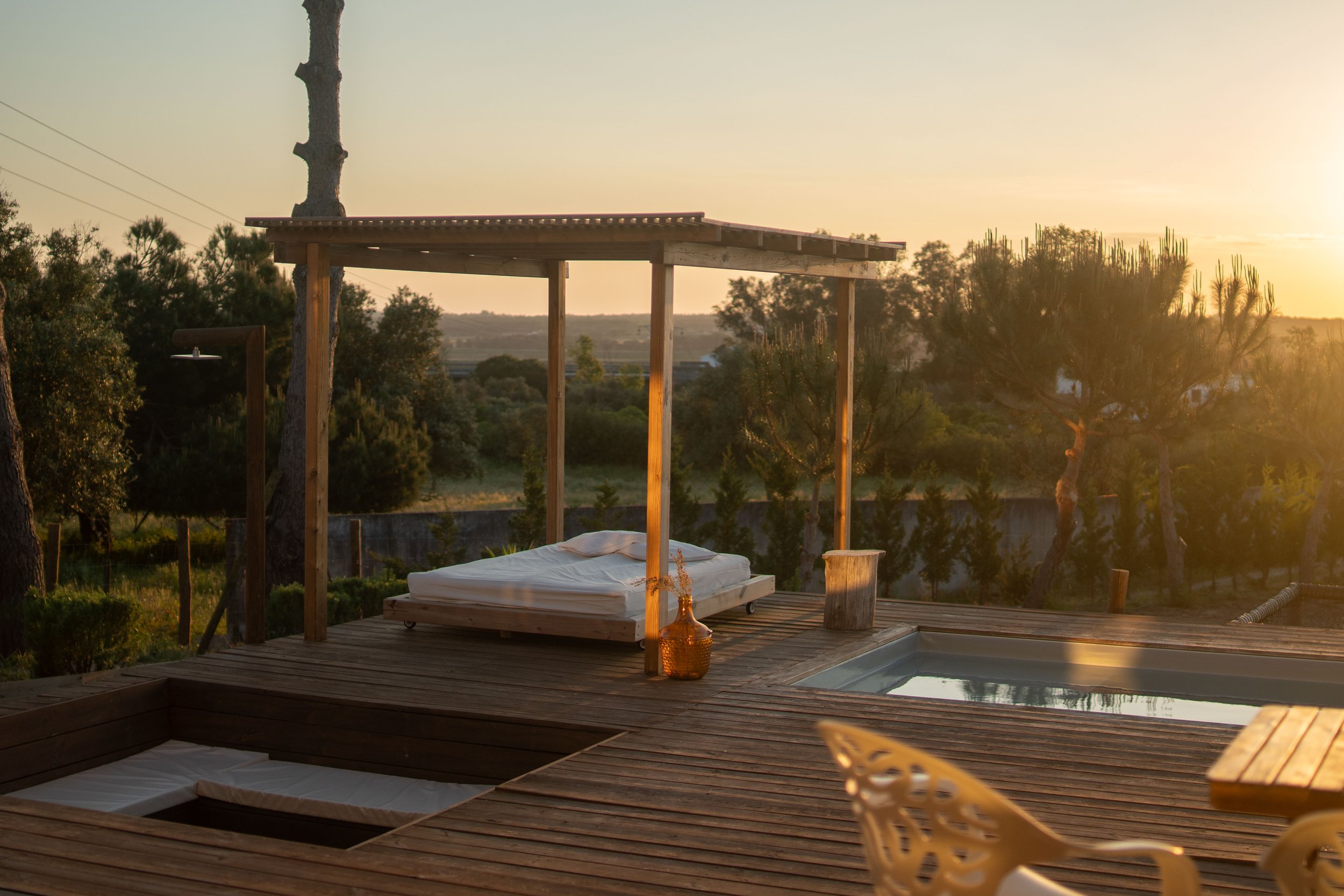Outdoor wooden deck with a bed, glass bottle, and a hot tub under a wooden pergola, surrounded by trees and shrubs during sunset.