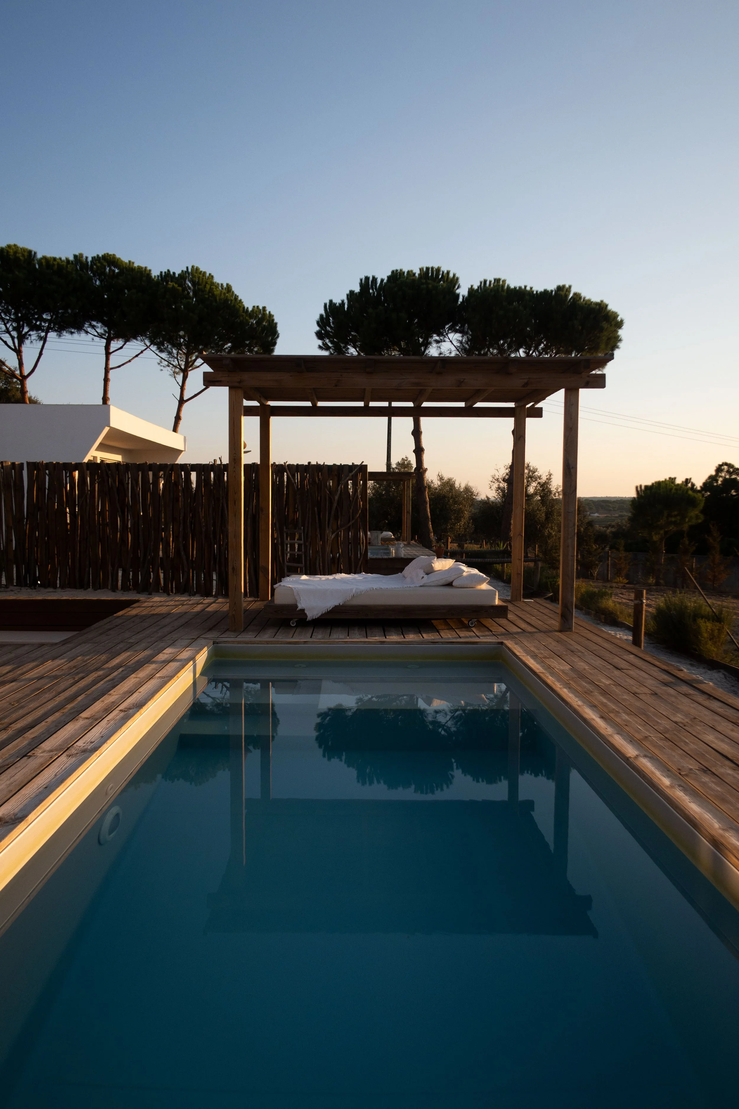 A modern backyard with a small swimming pool, wooden deck, and a covered lounge area with outdoor furniture and cushions, surrounded by a wooden fence and trees under a clear sky.