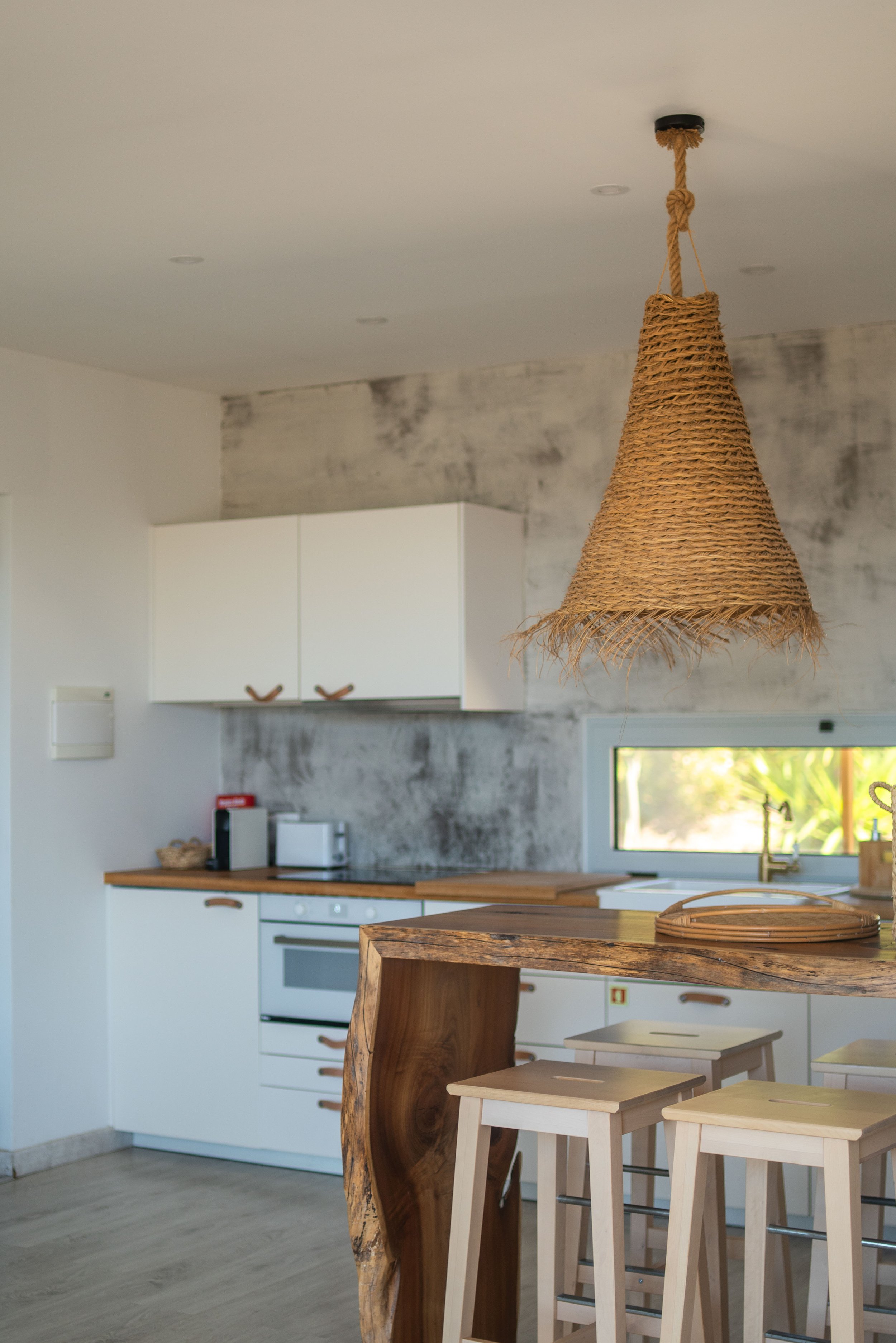 A modern kitchen with white cabinets, wooden countertops, a window, and a hanging wicker lampshade.