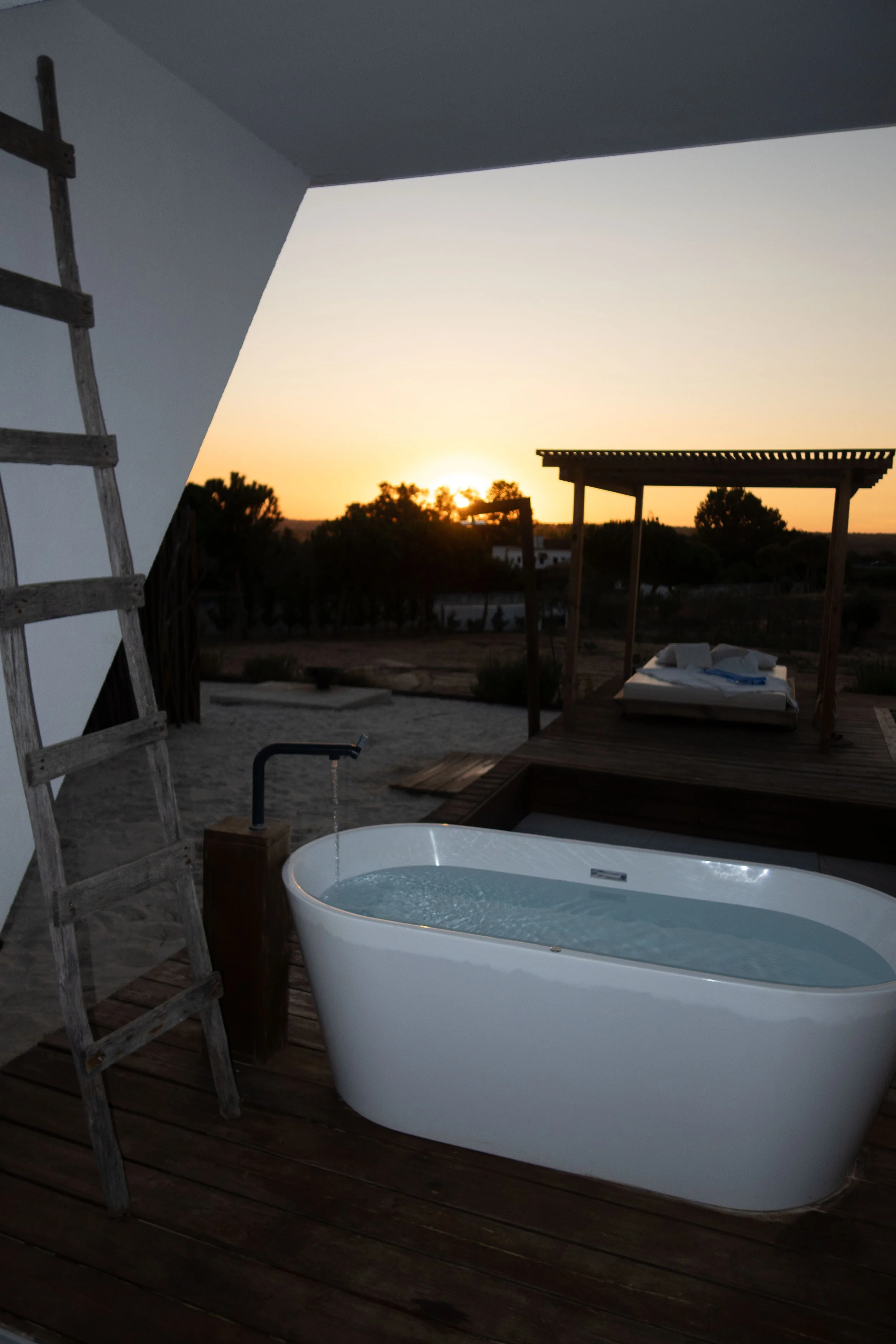 Outdoor scene at sunset with a bathtub filled with water on a wooden deck, a ladder on the left, and a wooden pergola on the right, with trees and buildings in the background.