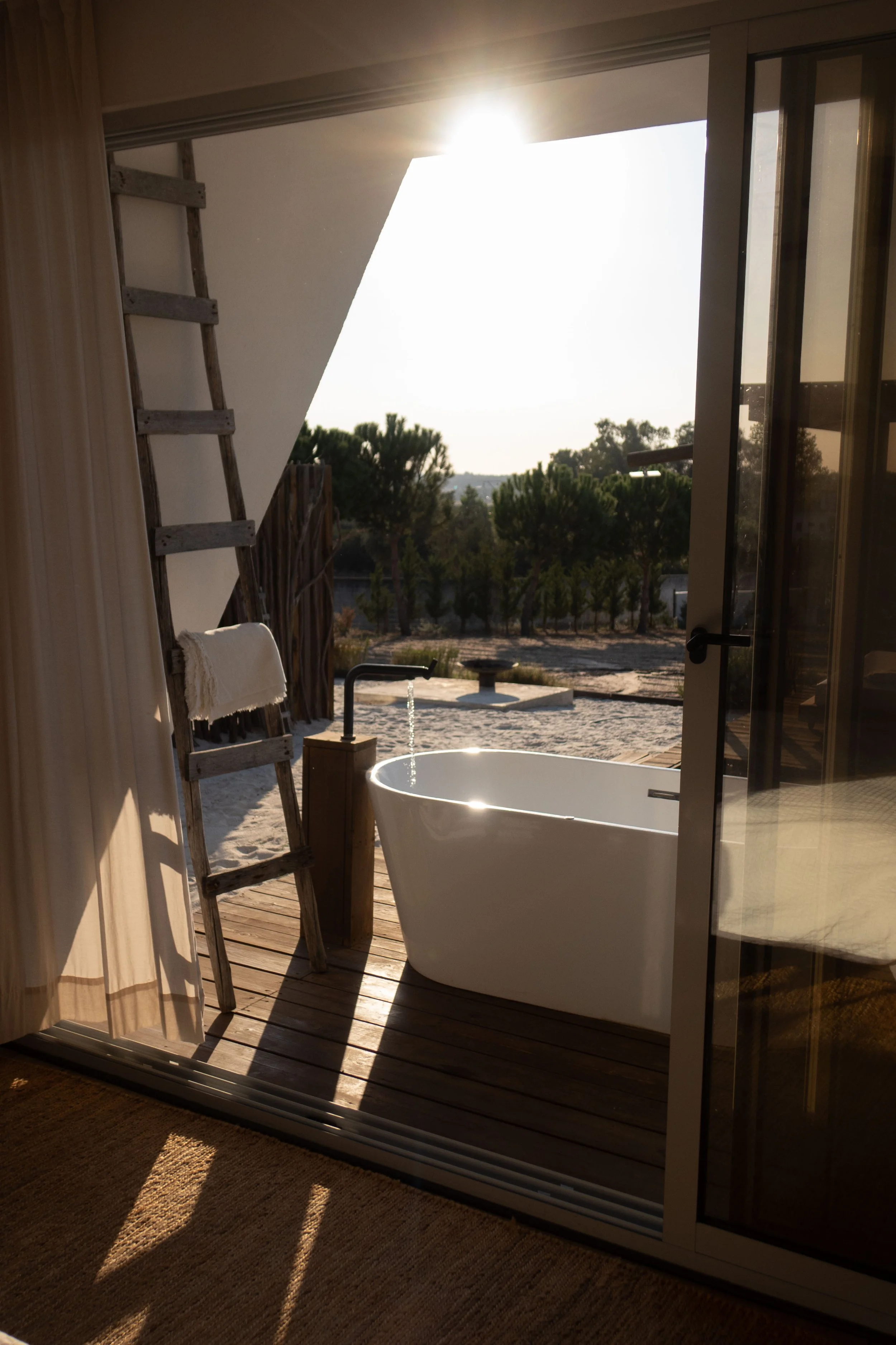 Sunset view through sliding glass door, showing a white bathtub on a wooden deck, with trees and sky in the background.