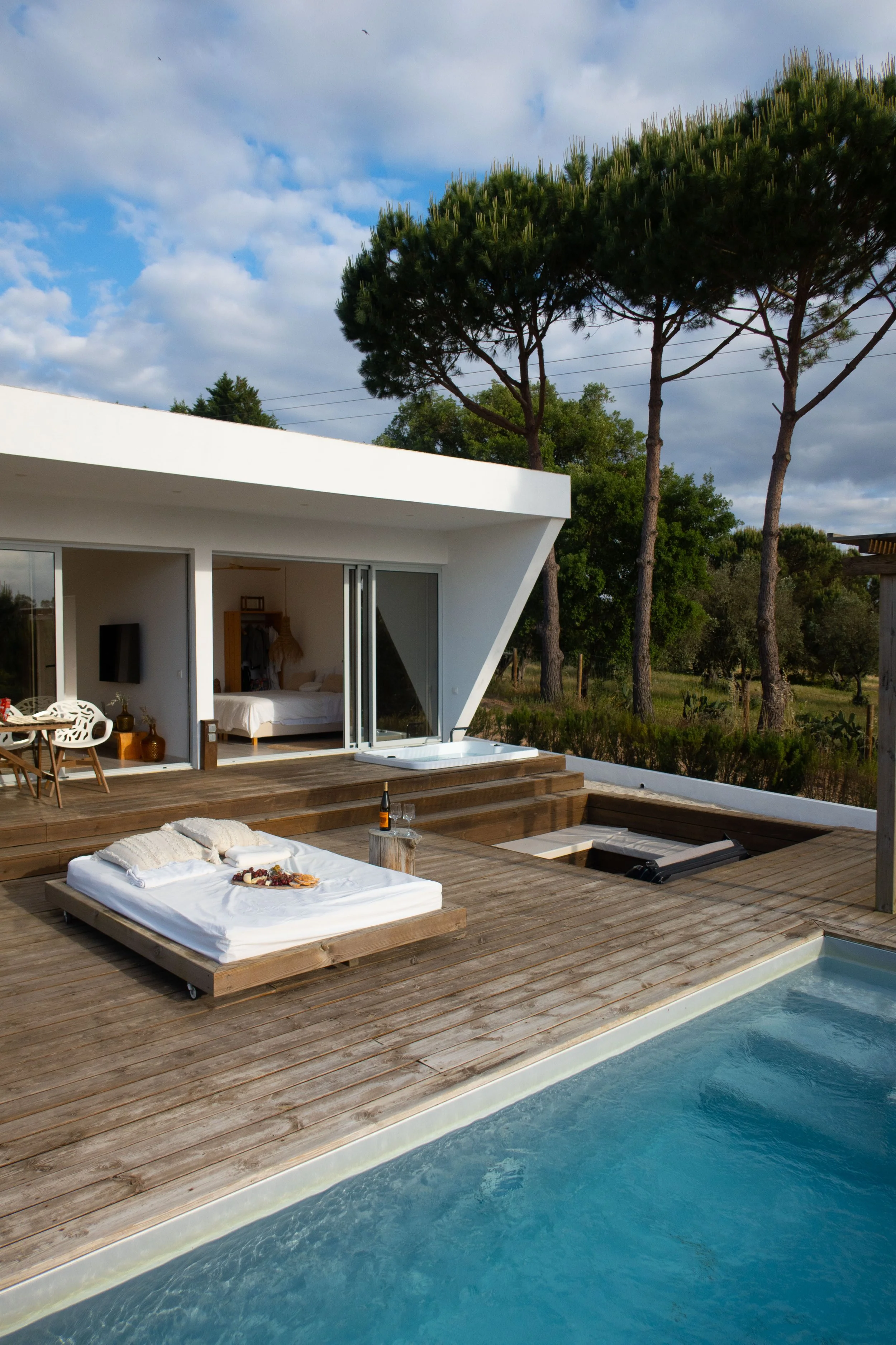 Modern house with large windows on wooden deck, overlooking a swimming pool, with outdoor lounging area, trees, and cloudy blue sky