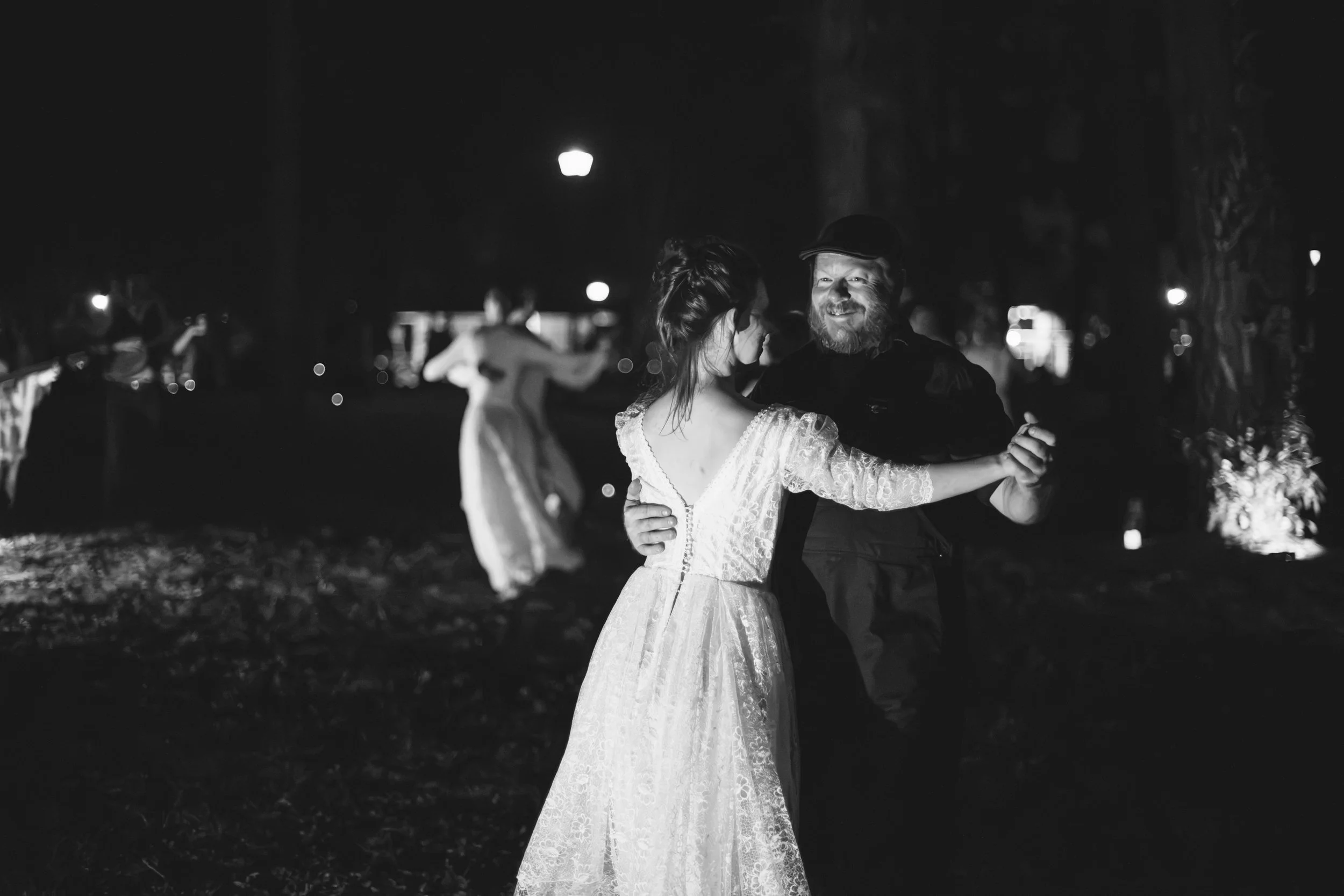 A black and white photo of a couple dancing outdoors at night. The woman wears a vintage lace dress and the man has a beard and cap. Other couples are in the background, also dancing.