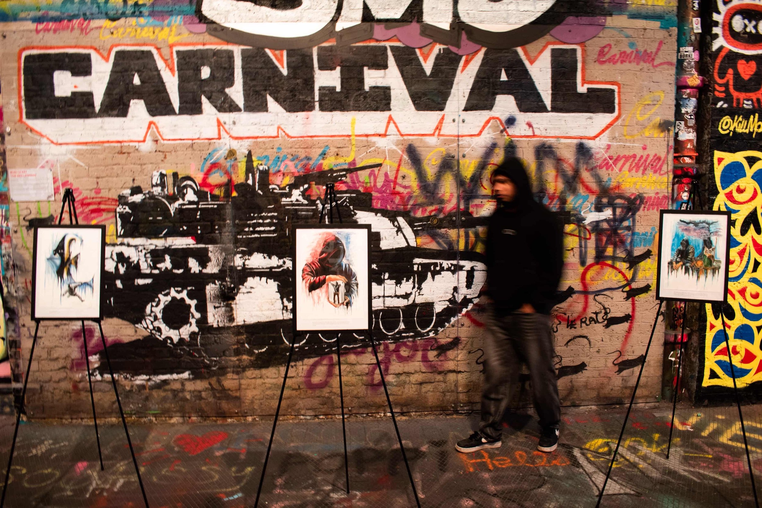 A brick wall covered in colorful graffiti art with a large black and white text reading 'ARREVIAL'. Several framed artworks are displayed on easels in front of the wall, and a person dressed in dark clothing is walking past. The floor is speckled with paint, and there are various stickers on the right side of the wall.