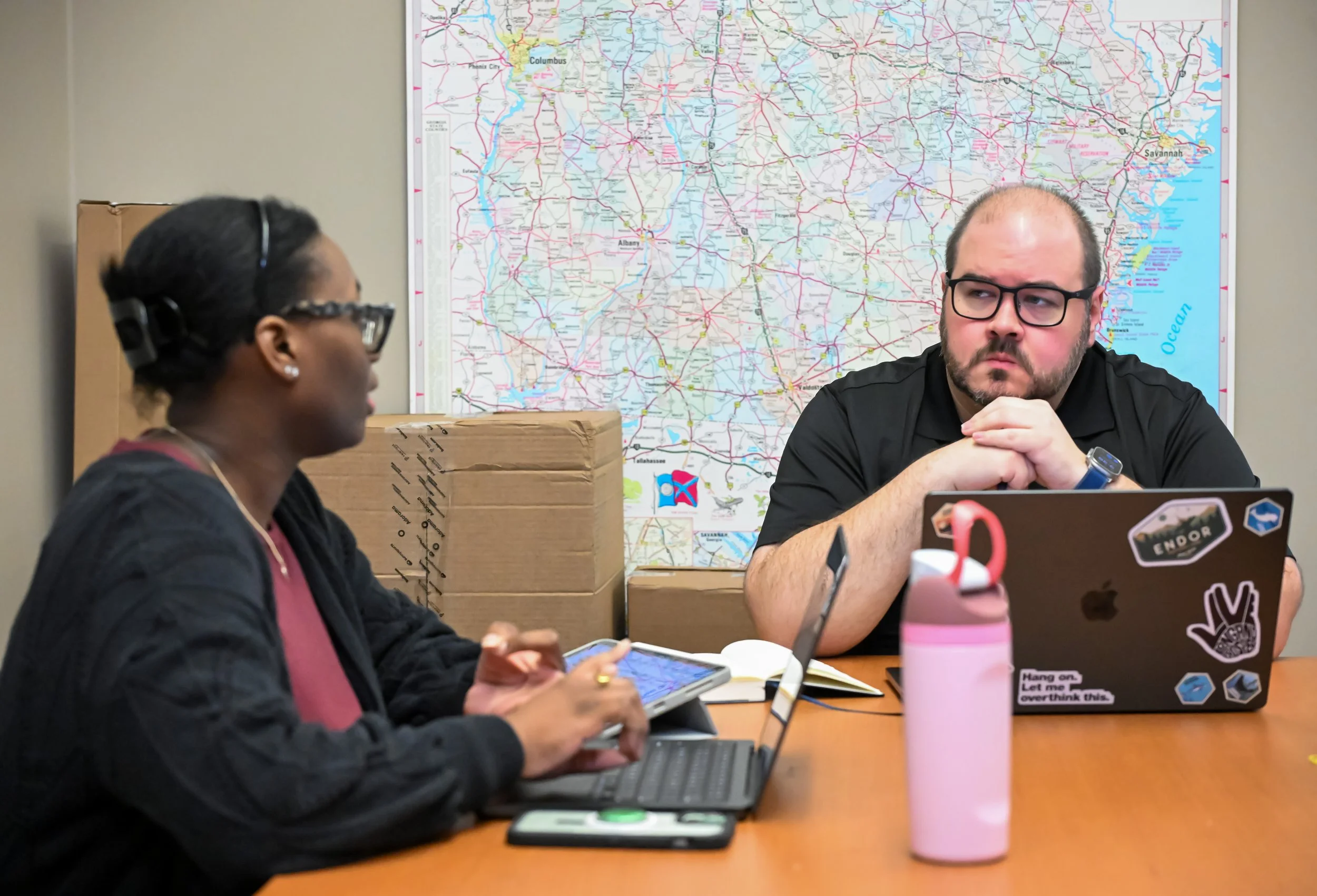 Two people seated at a table with laptops and tablets, in front of a large map of Georgia on the wall.