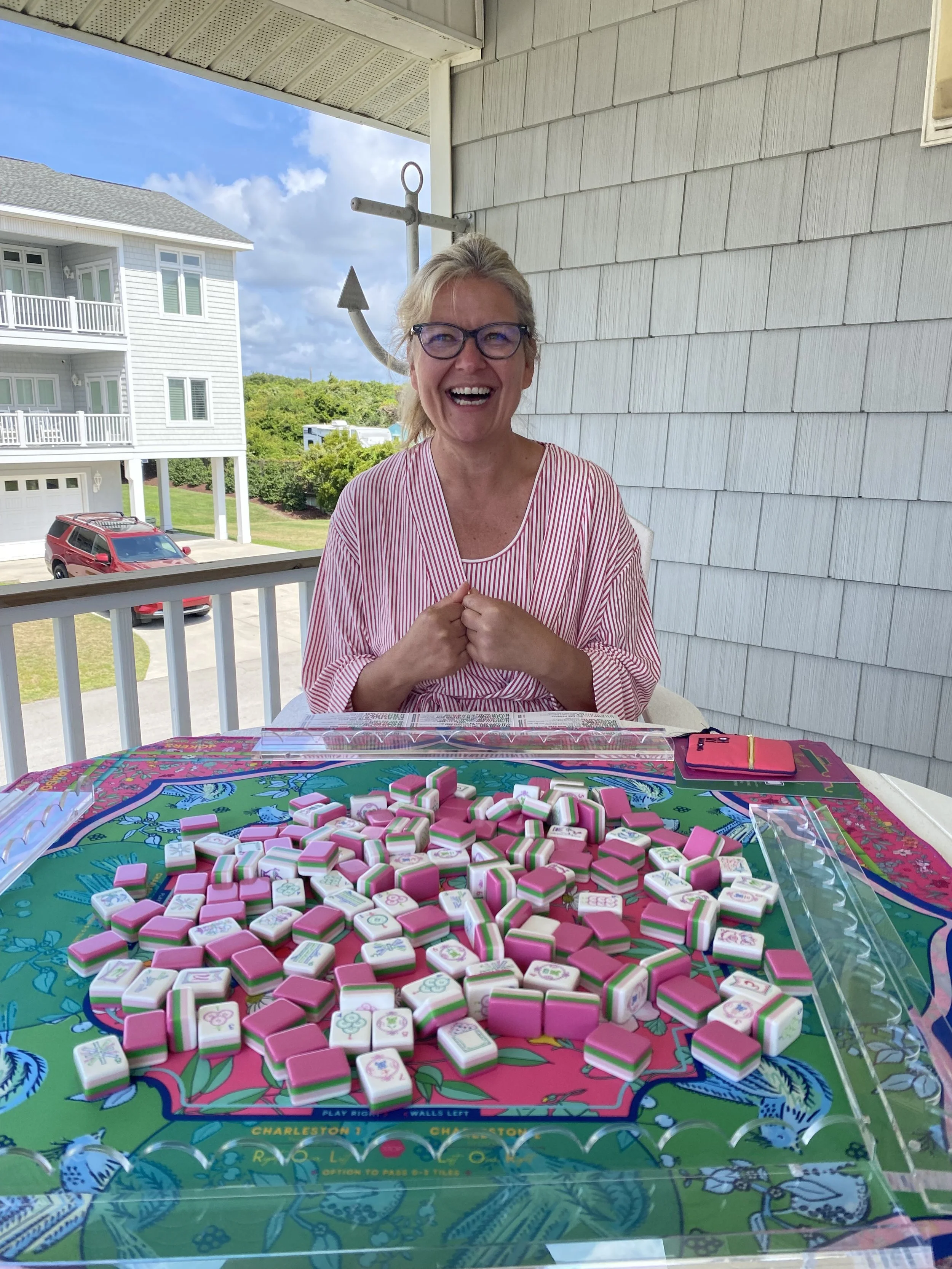 A woman with blonde hair, glasses, and a red and white striped shirt smiling and sitting at a table with a colorful mahjong game and pink and white tiles, outdoors on a balcony with a view of houses, trees, and a blue sky.