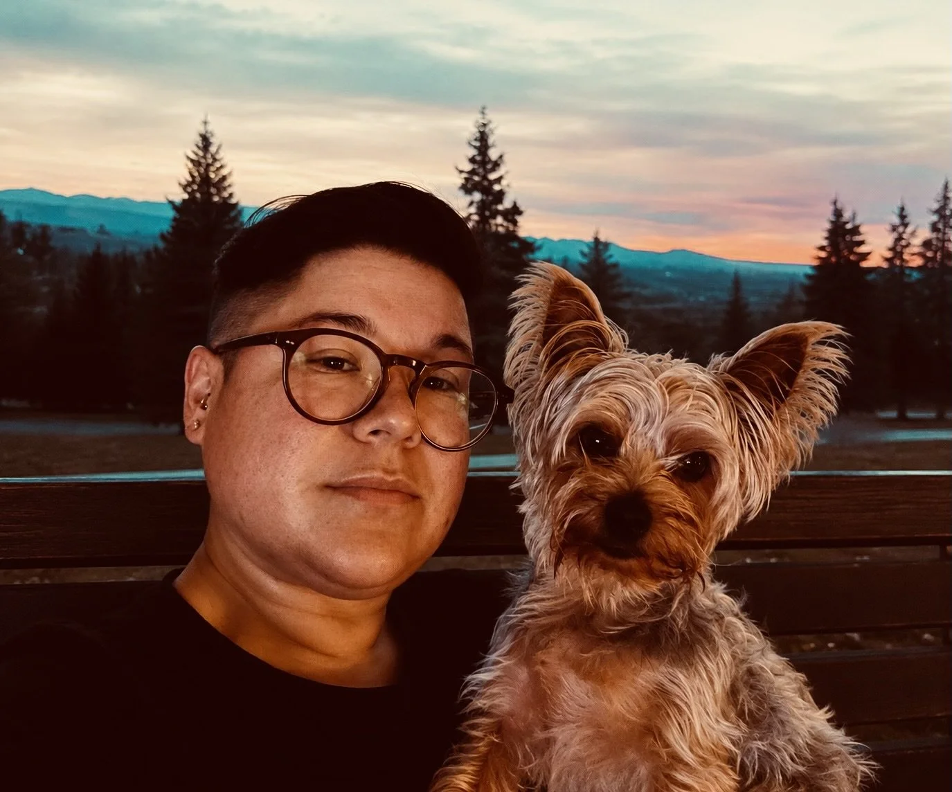 A person with dark hair, glasses, and earrings taking a selfie with a small dog with large ears, outdoors during sunset with trees and mountains in the background.
