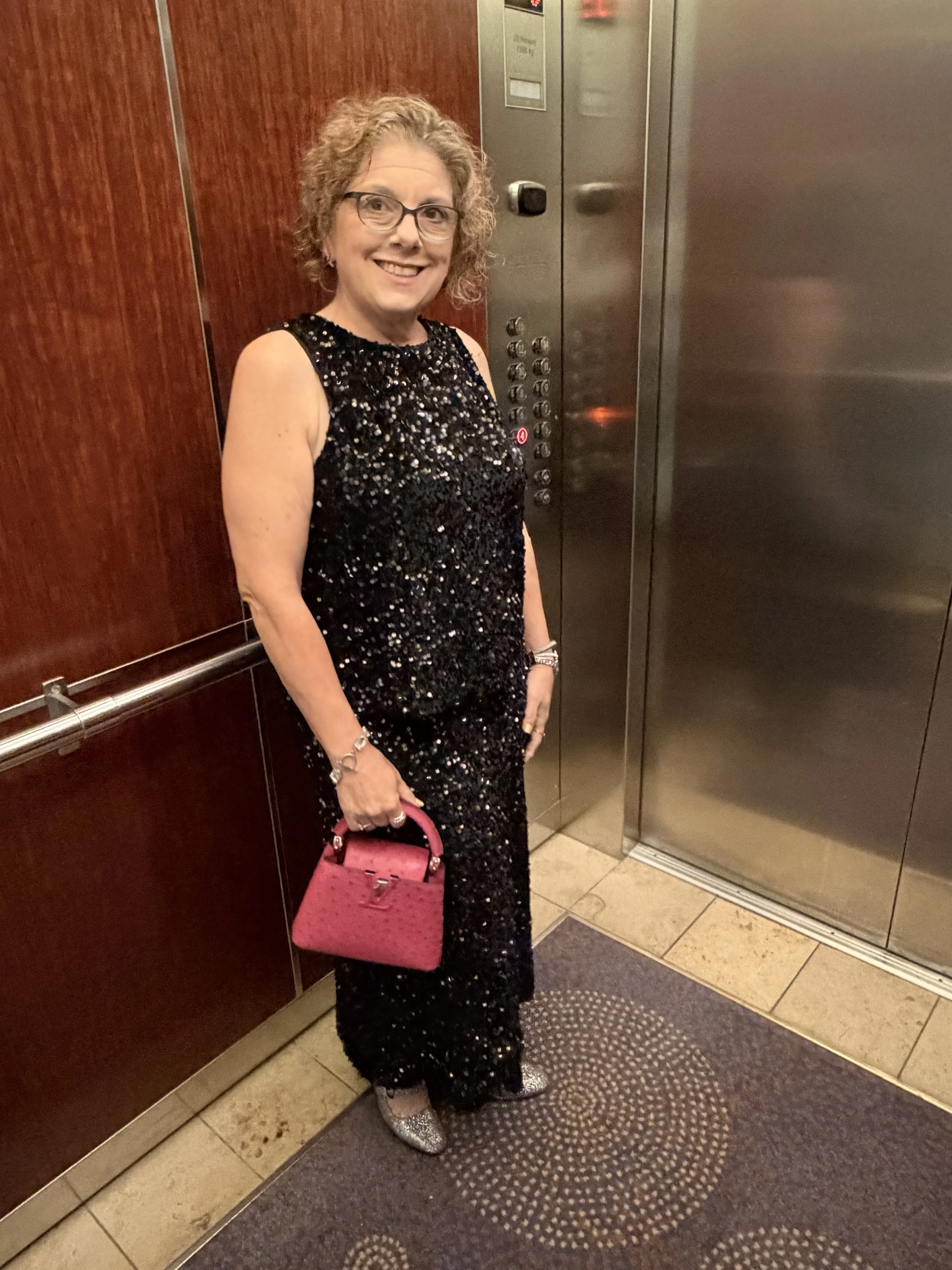 A woman in a black sequin dress holding a pink handbag inside an elevator lobby.