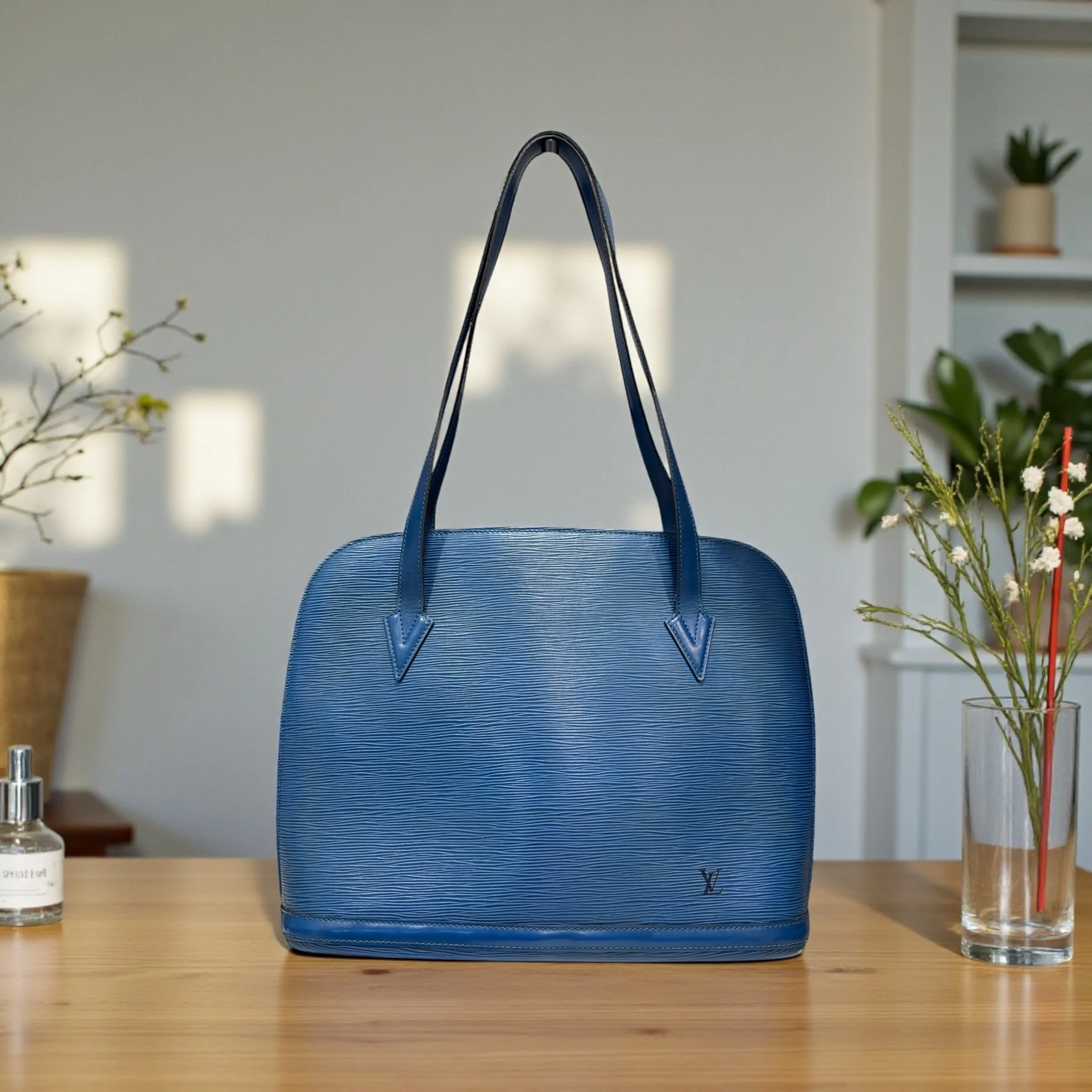 Close-up of a blue designer handbag on a wooden table, with two small bottles of skincare or cosmetic products on either side and green plants in the background.
