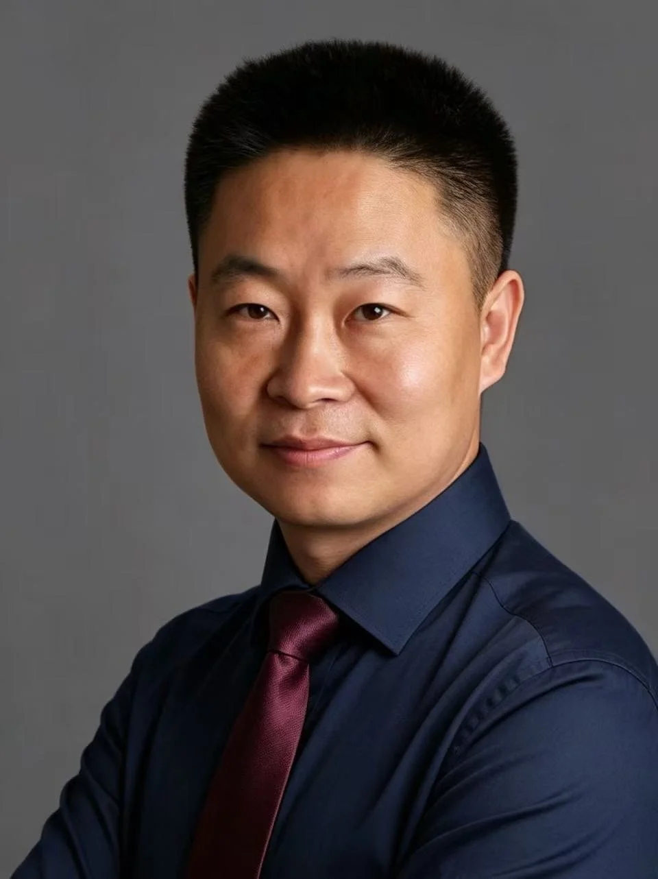 A professional headshot of an Asian man wearing a dark blue shirt and maroon tie against a gray background.