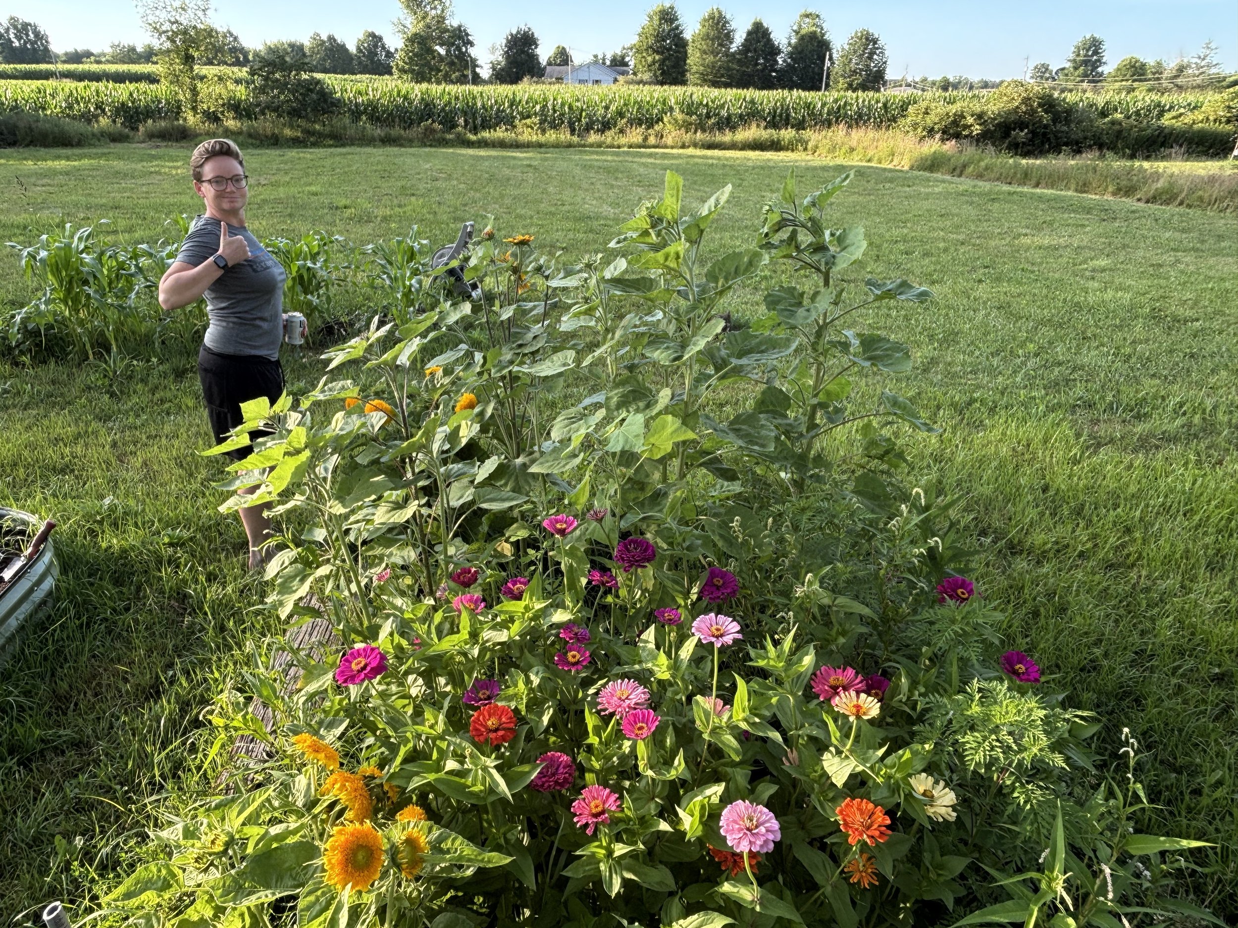 A person with glasses and short hair giving a thumbs-up, standing next to a flower garden with bright pink, orange, and yellow flowers, in a grassy field with rows of corn and trees in the background on a sunny day.