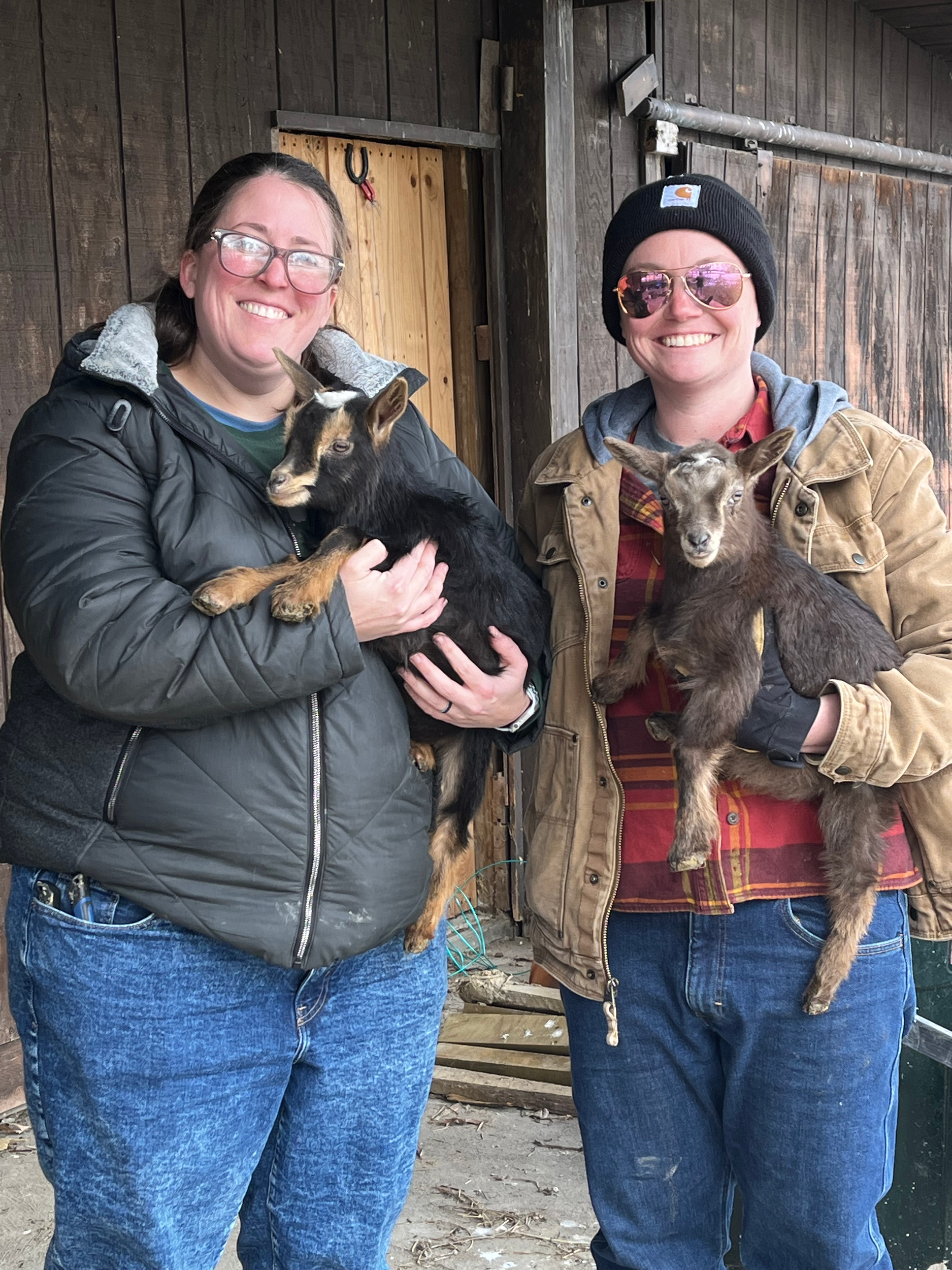 Two women standing in front of a wooden barn holding baby goats, one woman is wearing glasses, a black jacket, and blue jeans, and the other is wearing sunglasses, a black beanie, a brown jacket, and a red plaid shirt, with both smiling.