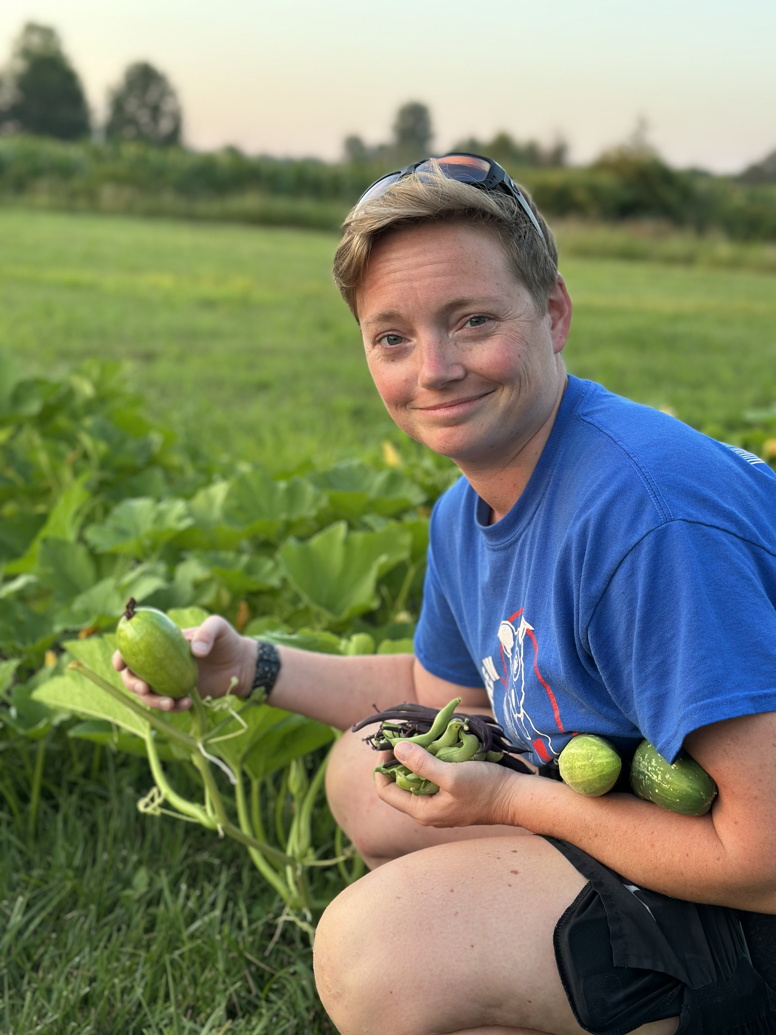 A person with short blonde hair, wearing a blue t-shirt and sunglasses on their head, kneels in a green field holding green vegetables and a small watermelon. The background shows a blurred nature landscape with trees and a clear sky.