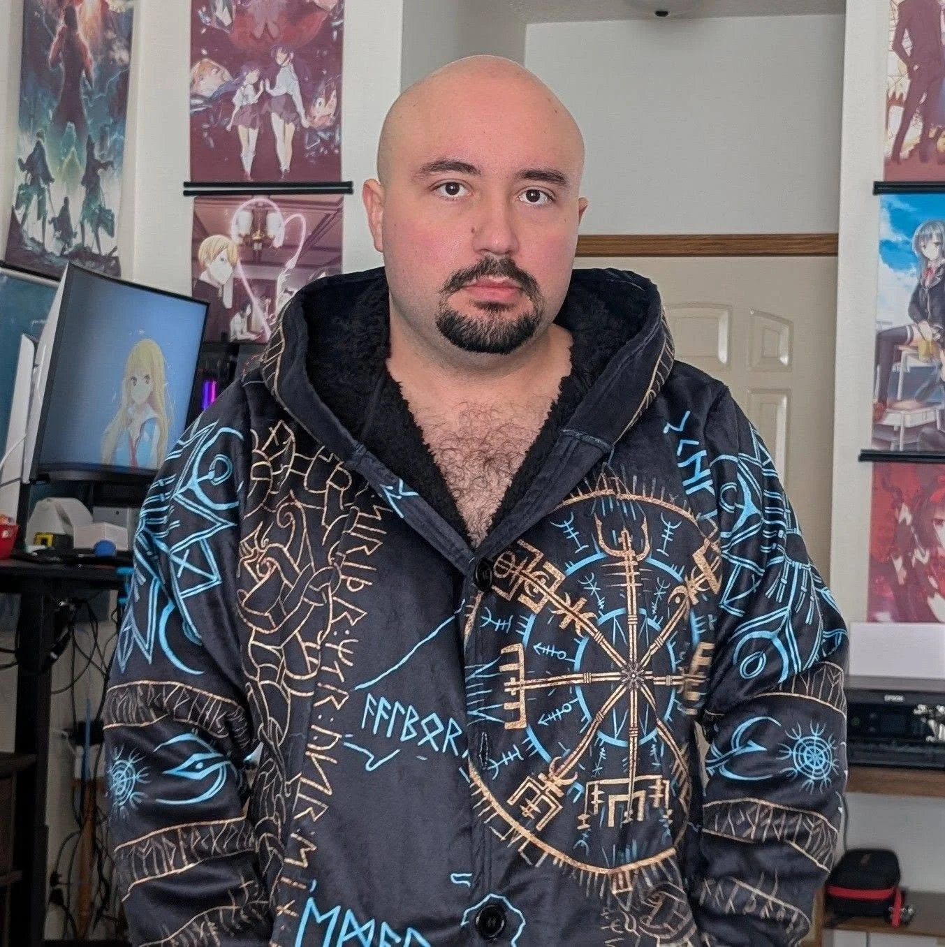A man with a makeup beard and mustache, wearing a black hoodie, sitting in a room with anime posters on the walls and computer monitors.