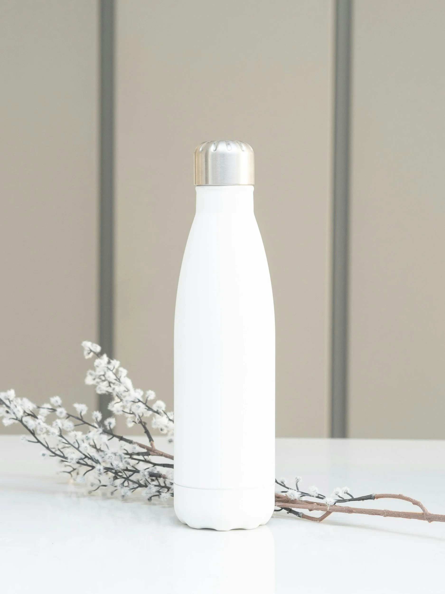 A white soap or lotion dispenser with a metallic cap, placed on a white surface. Behind it is a branch with white blossoms and a beige background with vertical grey lines.