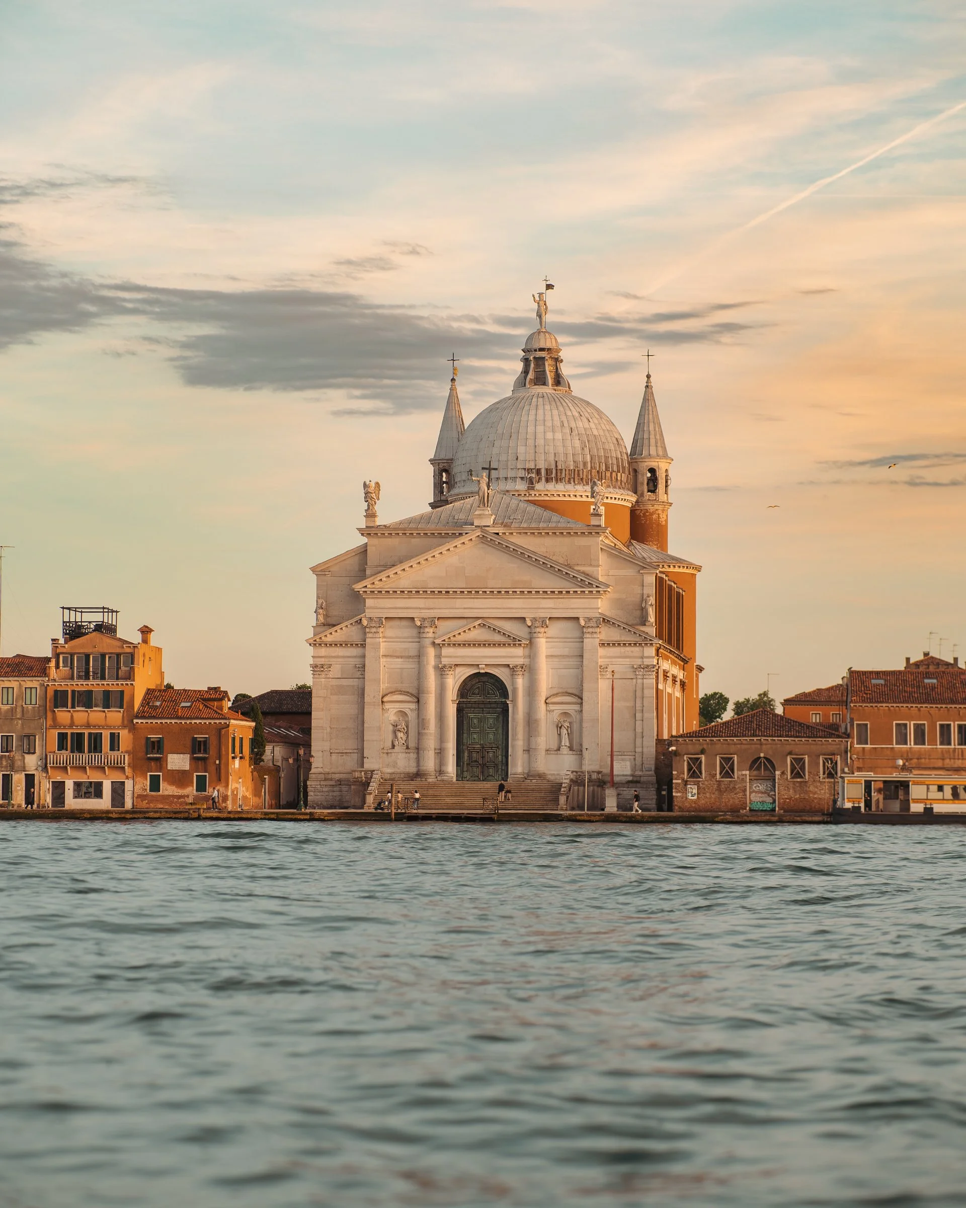 A historic church with a large central dome, flanked by four smaller towers, seen from a venitian canal at sunset with soft orange and blue sky, and buildings along the shoreline.