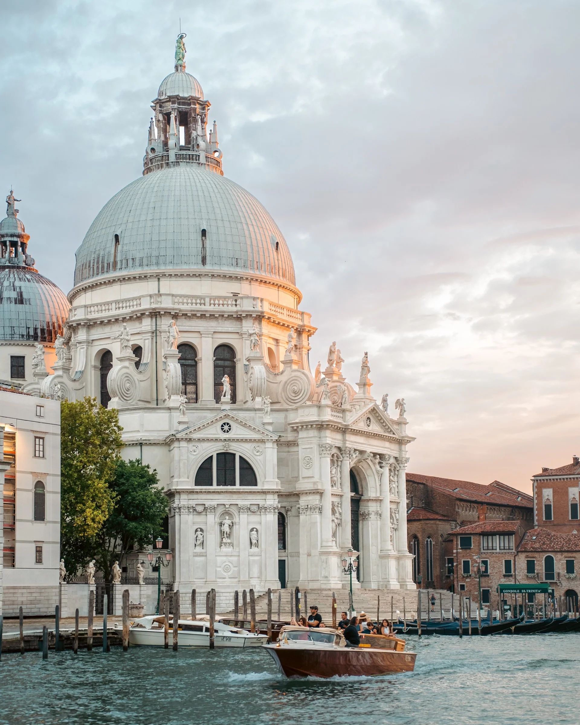 View of a white ornate church in Venice, with multiple domes and statues along the waterfront, with boats and gondolas on the canal, and people walking along the promenade.