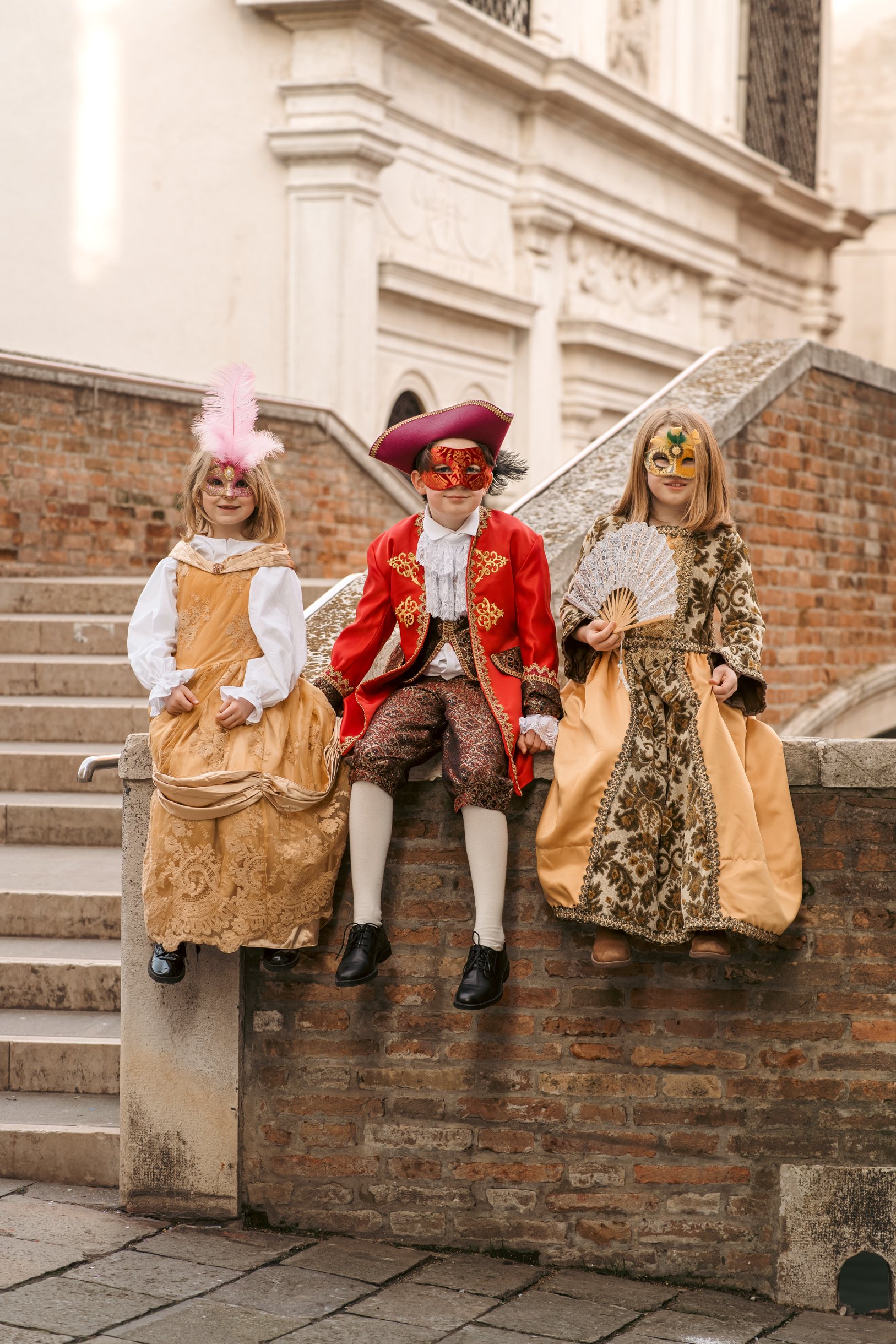 Three children dressed in carnival costumes are standing on a brick and concrete staircase outdoors, in front of a historic building, in Venice, Italy.