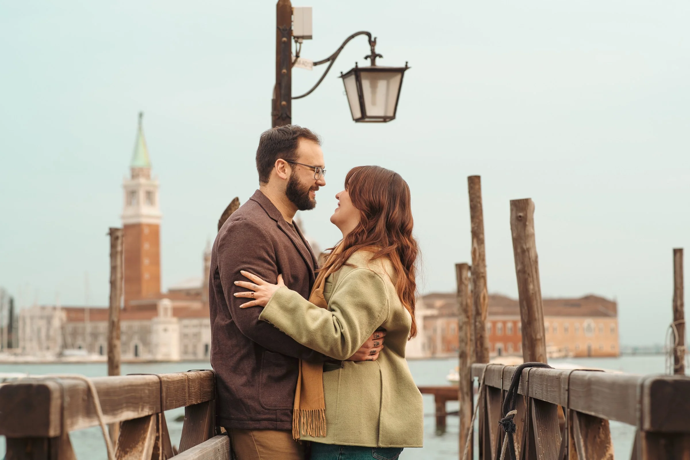 A couple is standing on a wooden dock, embracing and smiling at each other, in Venice, Italy.