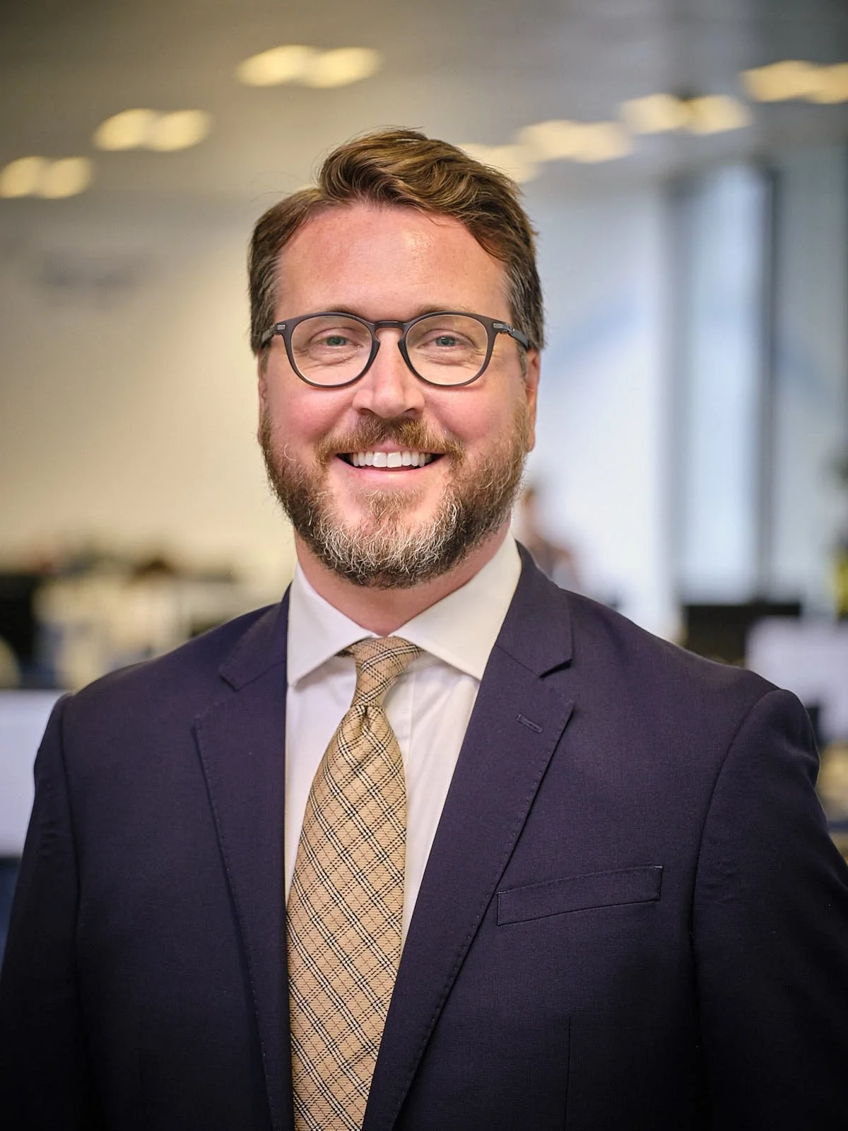 CEO of VentureTech Partners 'Mark McKenna' smiling with glasses, beard, and brown hair in a professional office setting, wearing a navy suit, white shirt, and patterned beige tie.