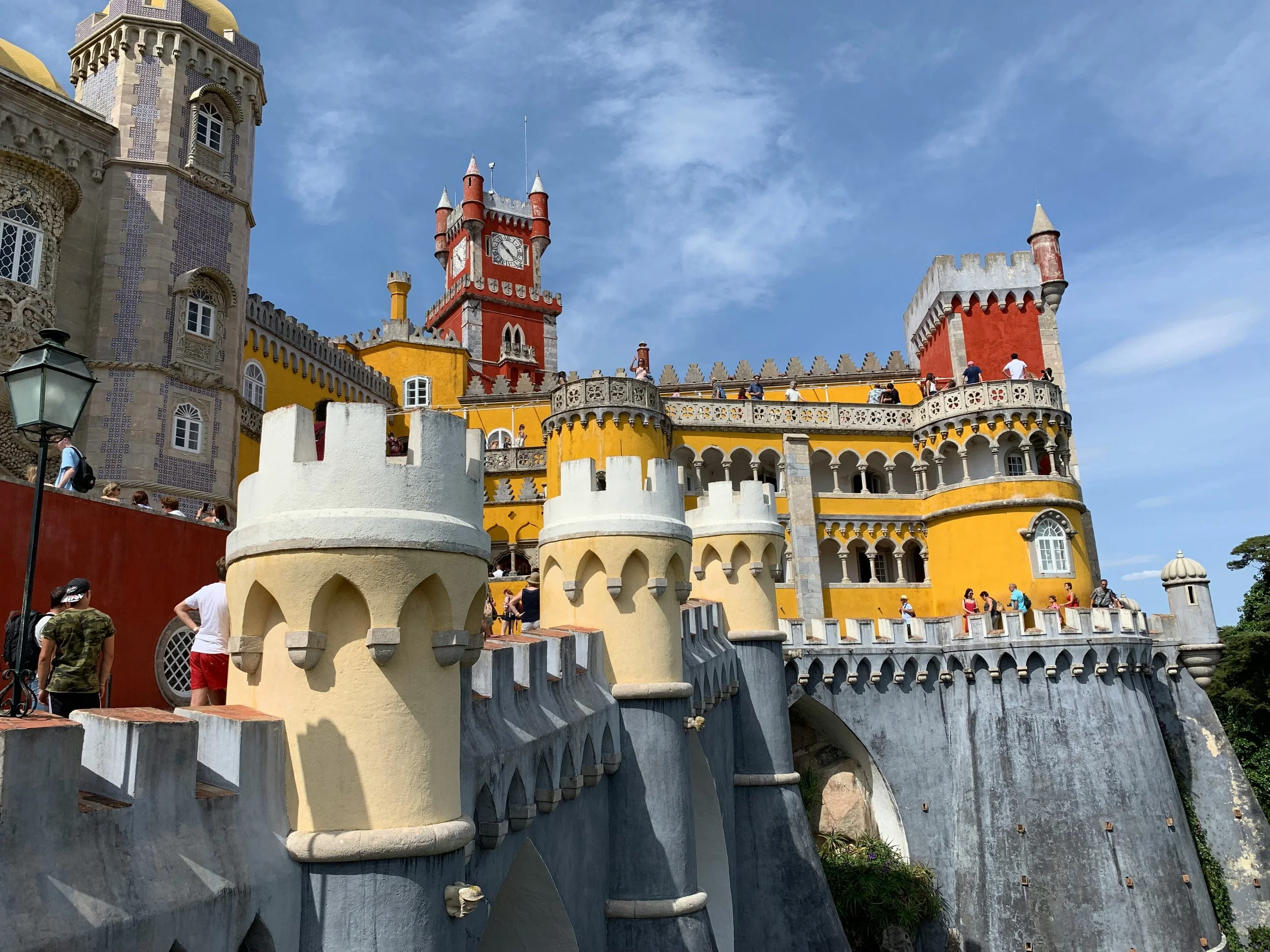 Colorful fairytale castle with yellow, red, and beige turrets and towers under a bright blue sky with some clouds, with visitors walking around.
