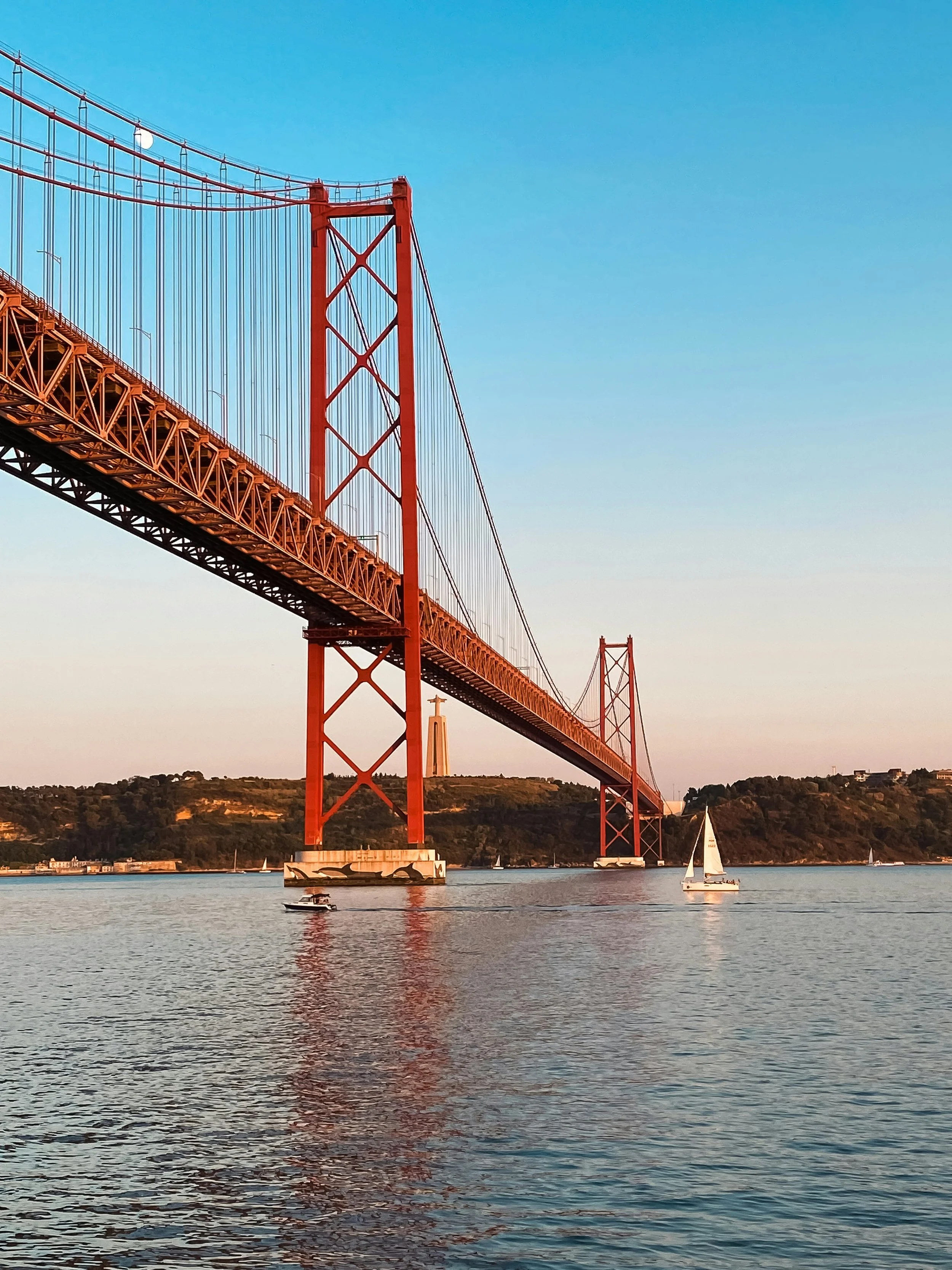 The image shows the Golden Gate Bridge in San Francisco, California, with sailboats and a motorboat on the water, and green hills in the background under a clear blue sky.