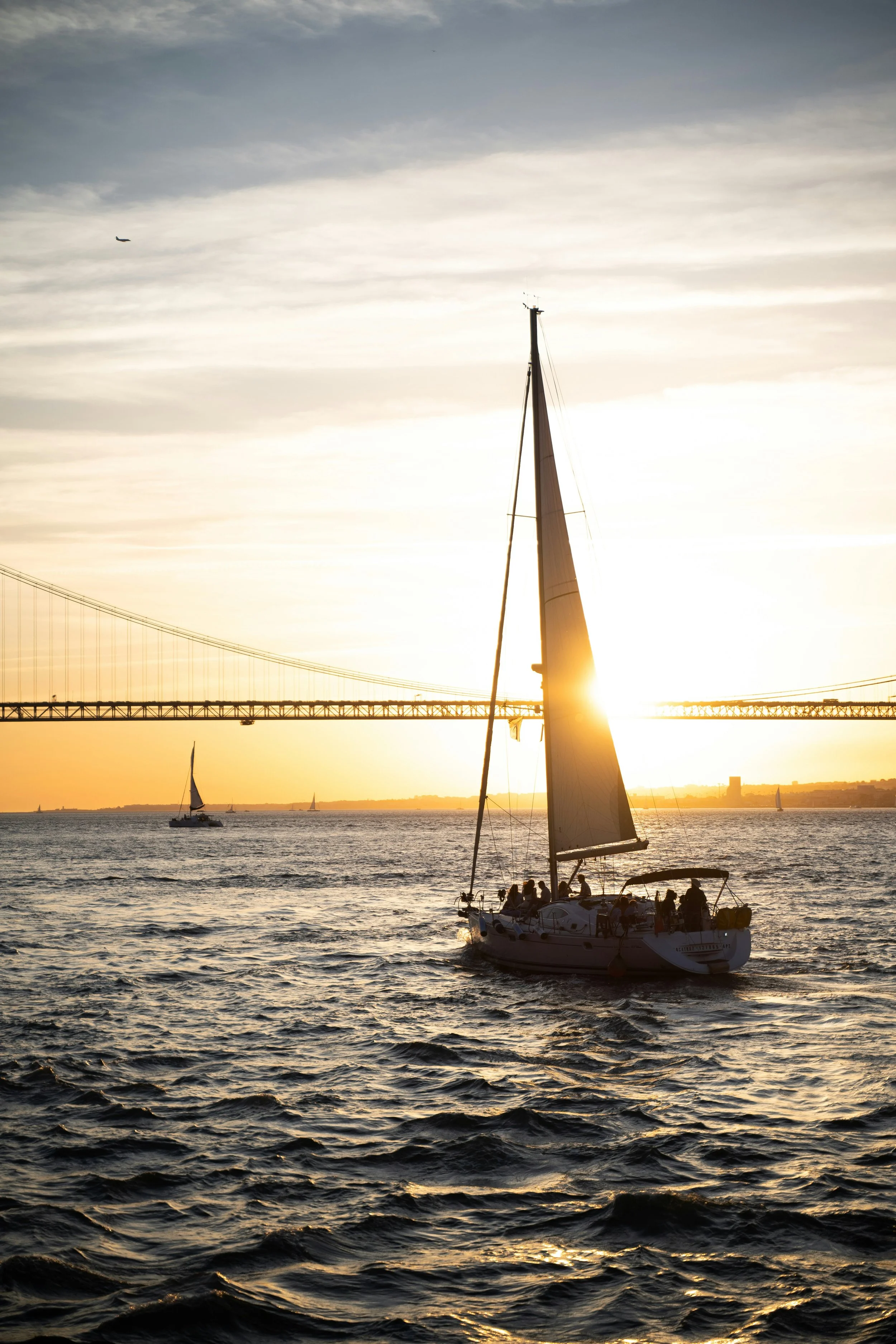 A sailboat with passengers on the water during sunset, under a sky with some cloud cover and a distant bridge.