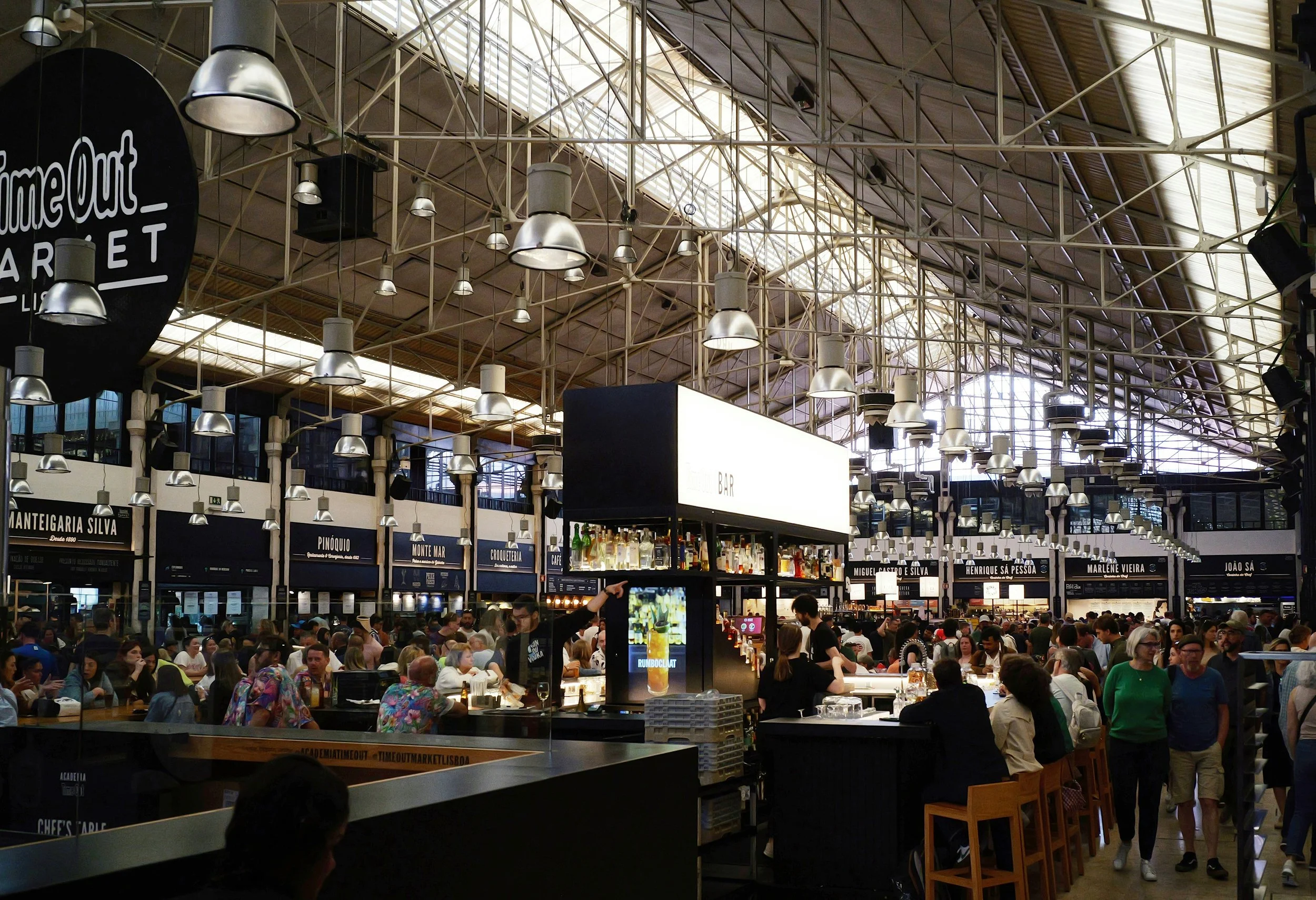 A crowded indoor market with high ceilings, metal framework, and numerous hanging lights. The market has vendor stalls selling various goods, and many people are shopping and walking around.