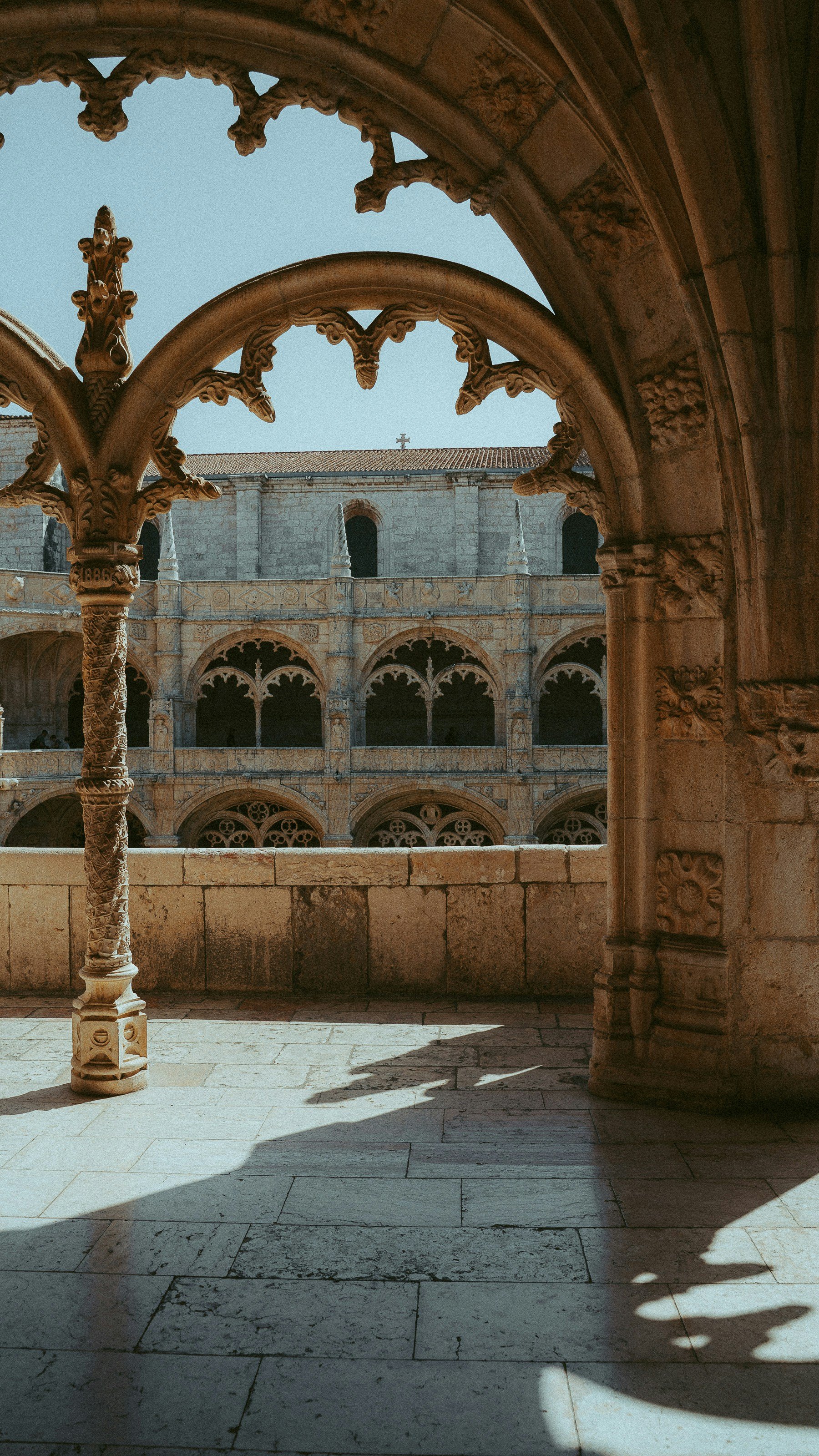 View of a historic stone courtyard with ornate arches and a stone building with a cross on the roof in the background, seen through a carved stone archway.
