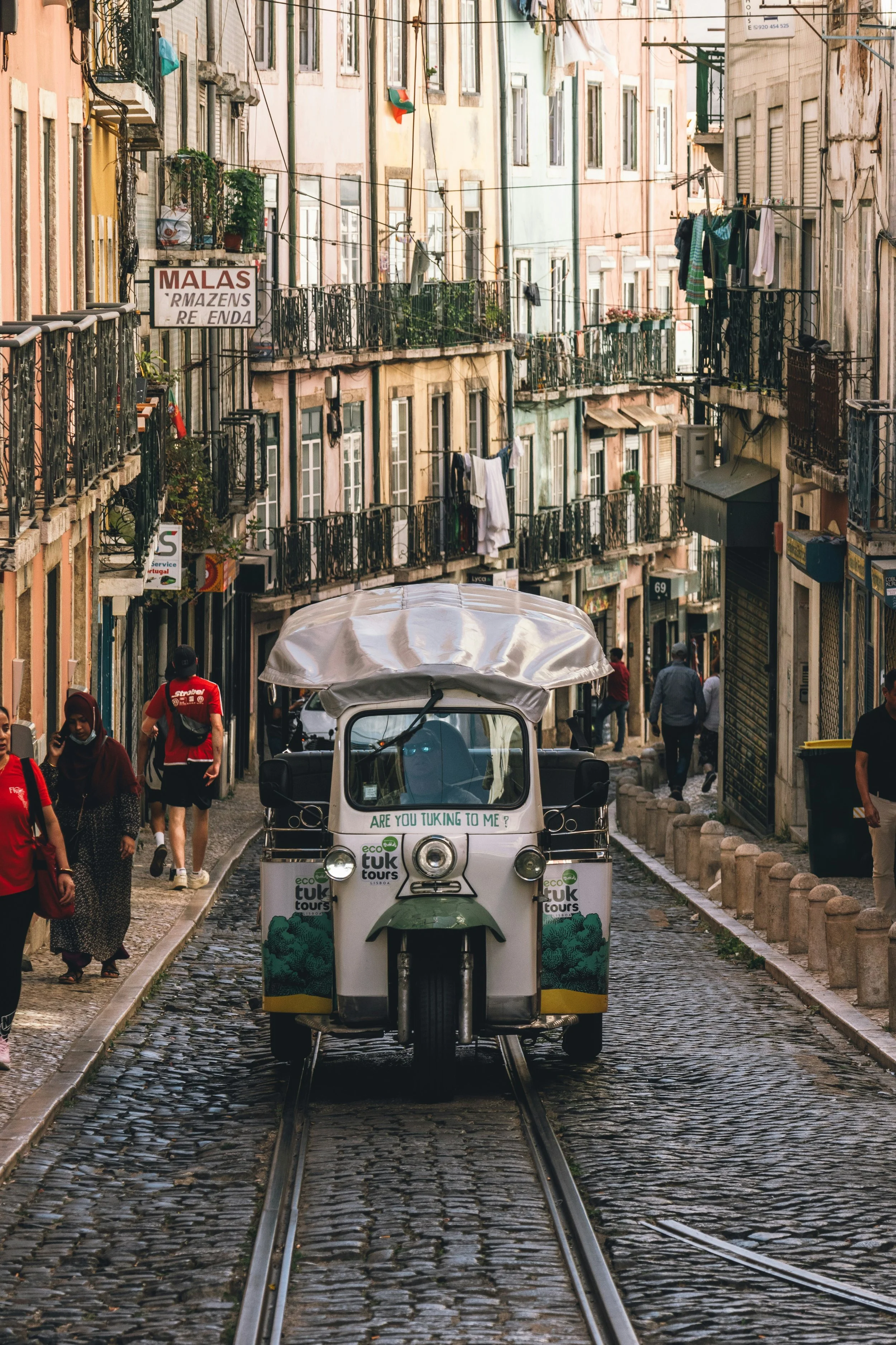 A street scene in Lisbon, Portugal featuring a small electric tuk-tuk on cobblestone tracks with pedestrians walking along the sides. The background displays colorful buildings with small balconies and laundry hanging out to dry.